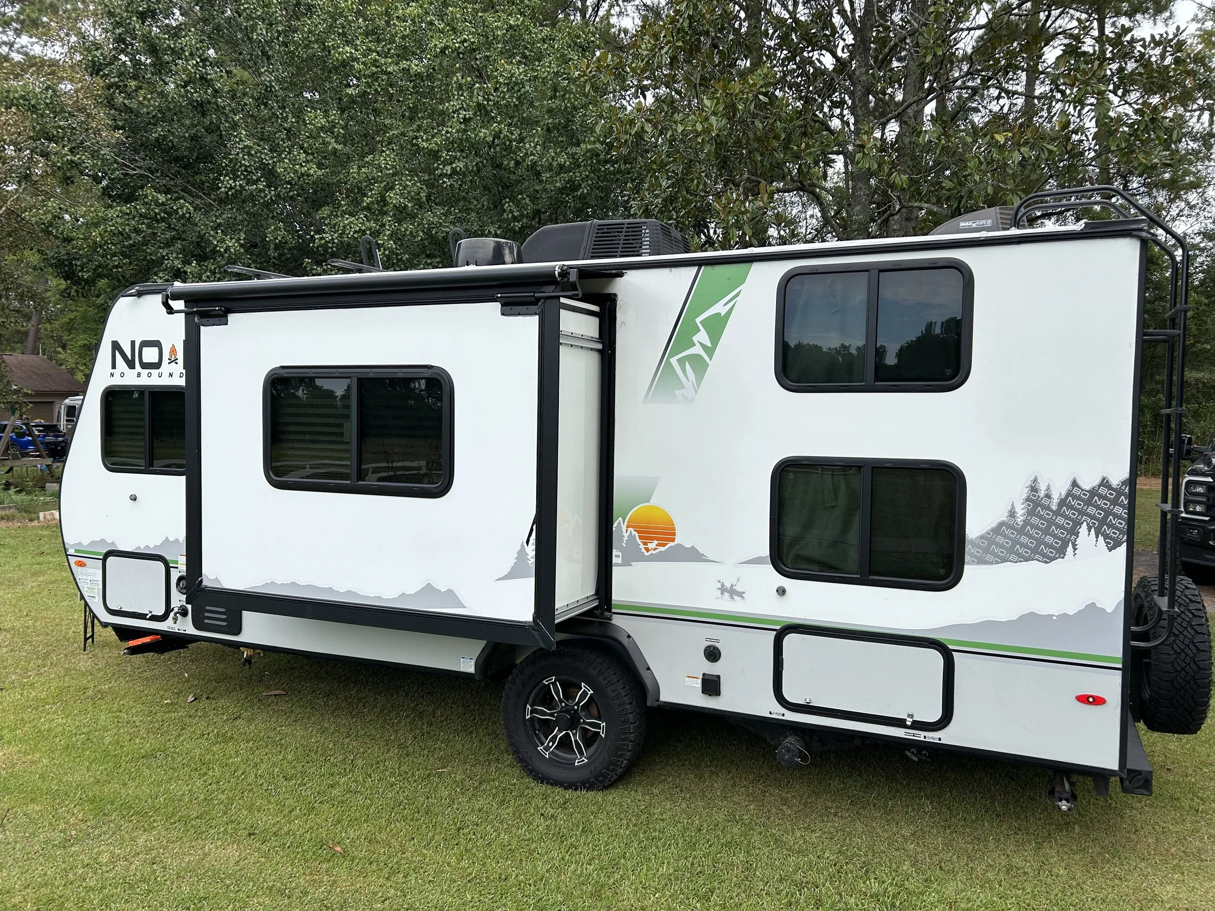 A modern white travel trailer with multiple windows, a fold-out slide, and mountain-themed graphics on the side, parked on a grassy area with trees in the background.