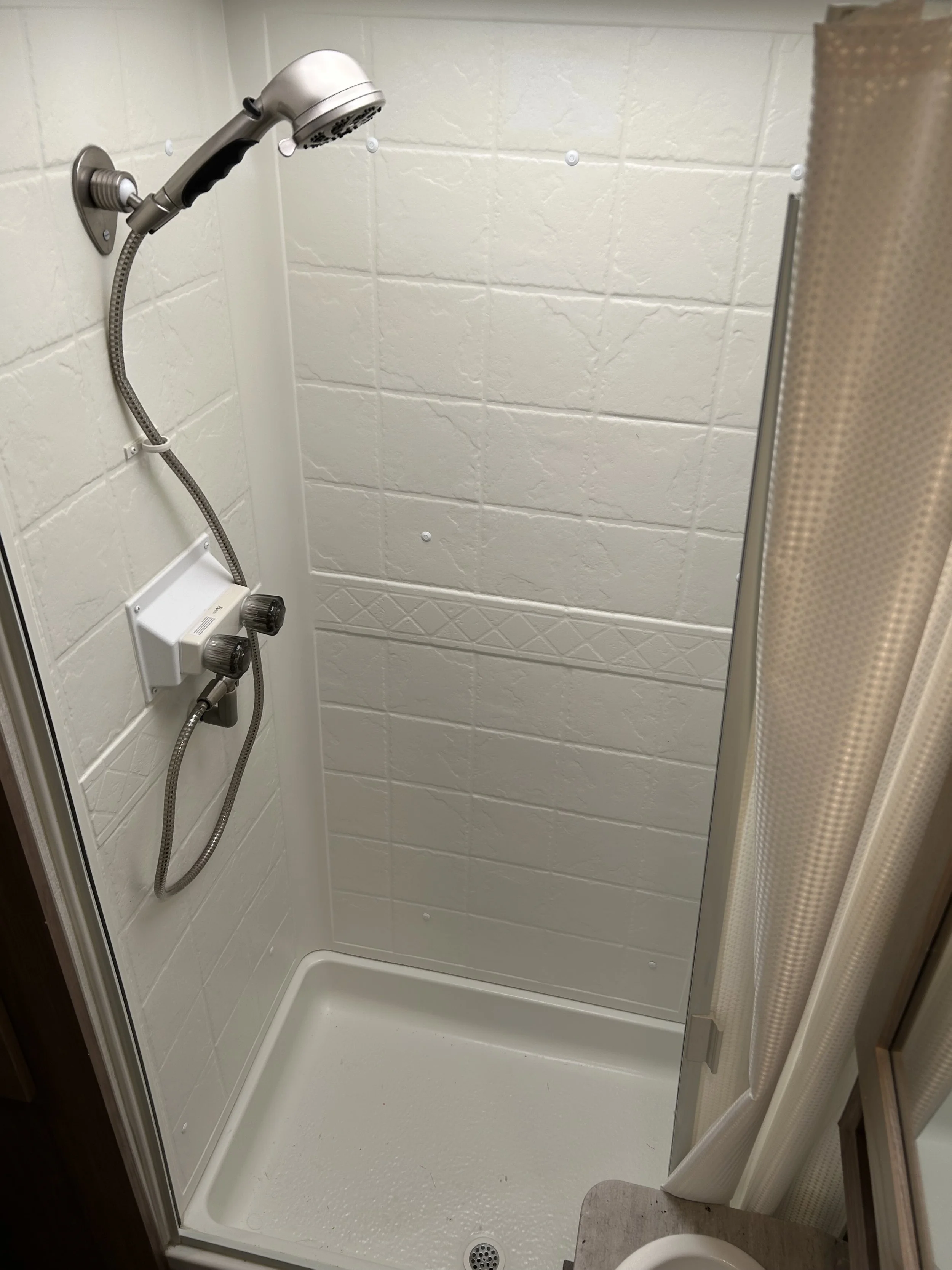 Compact shower stall with white tiled walls, a silver handheld showerhead, and a white shower base. Part of a beige shower curtain and a bathroom fixture are visible.