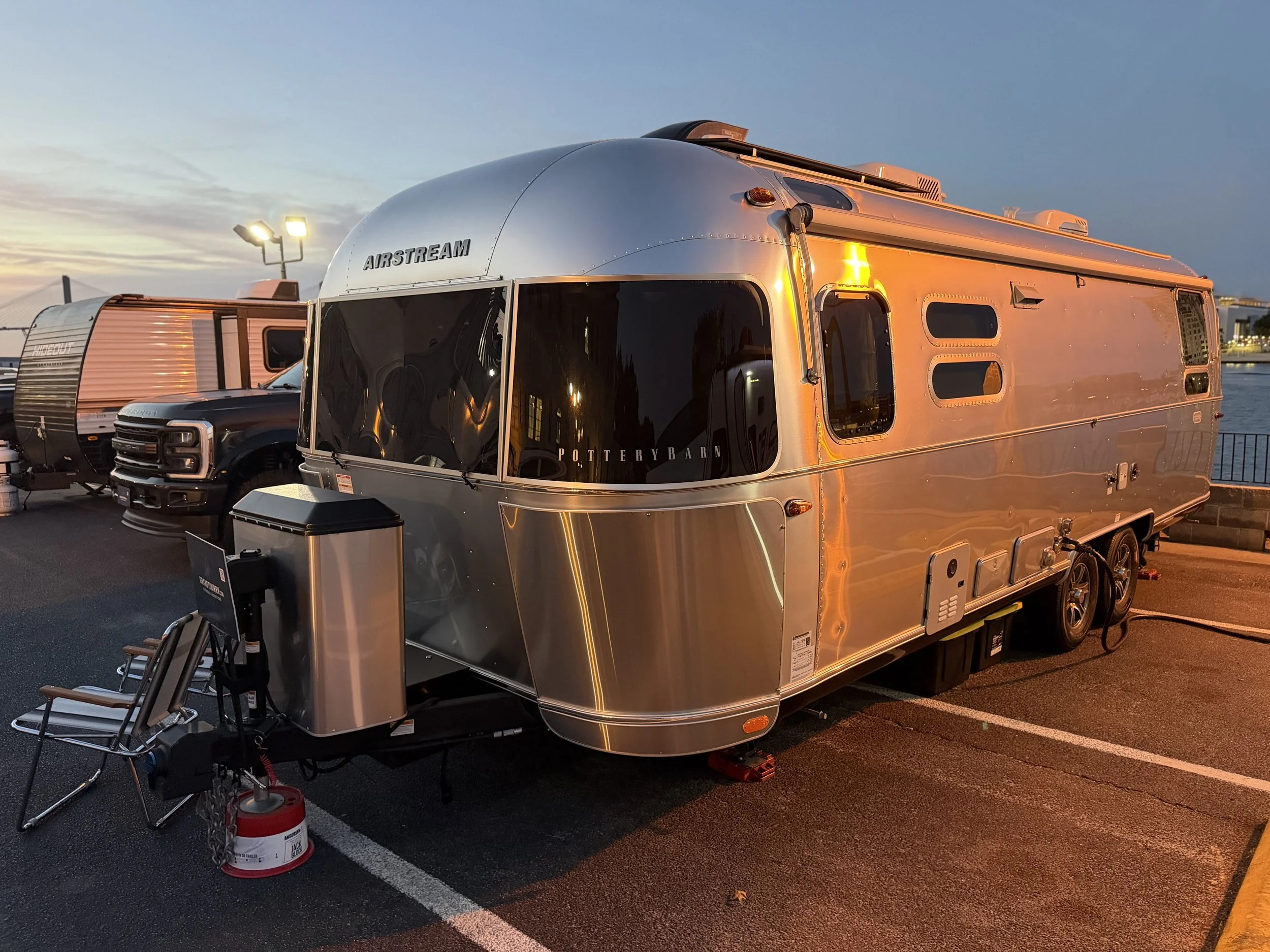 A silver Airstream travel trailer parked in a lot during dusk, with adjacent vehicles and water visible in the background.