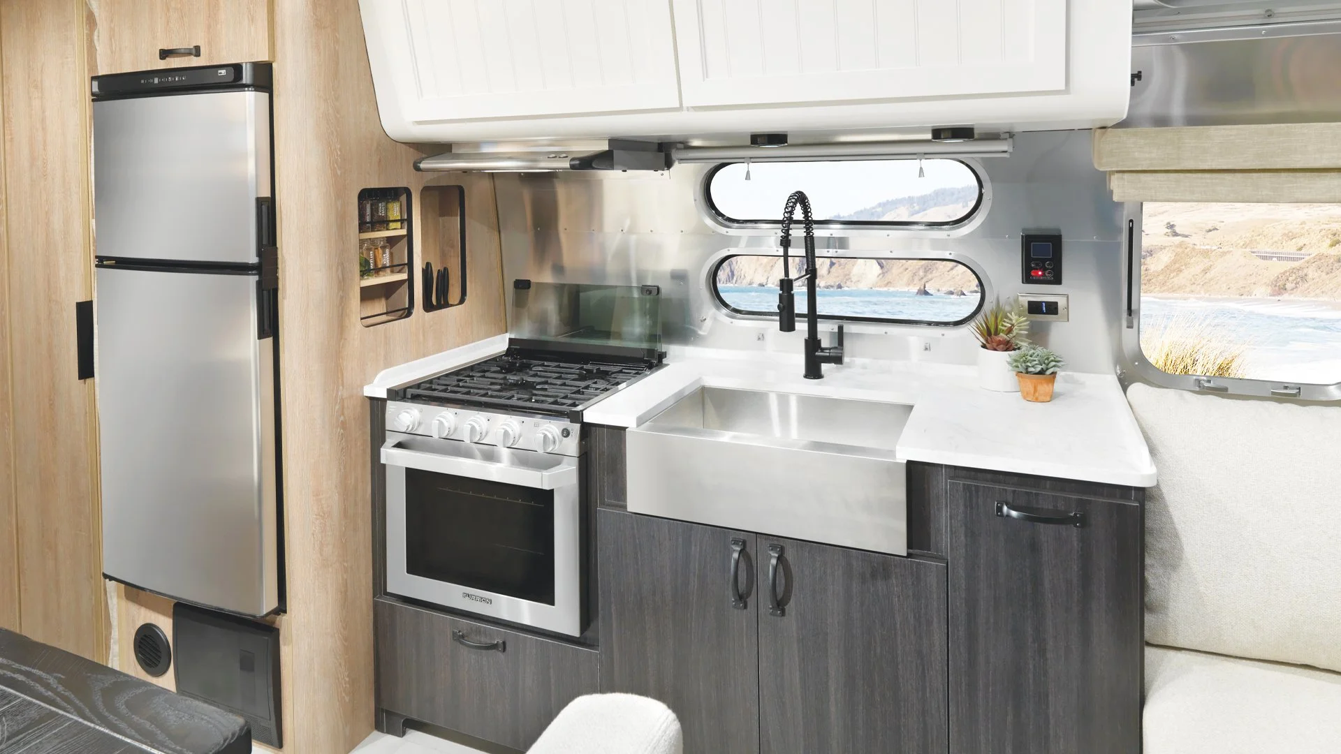 Small kitchen area inside a camper or RV with a refrigerator, stove, sink, and cabinets, overlooking a river view through rounded windows.