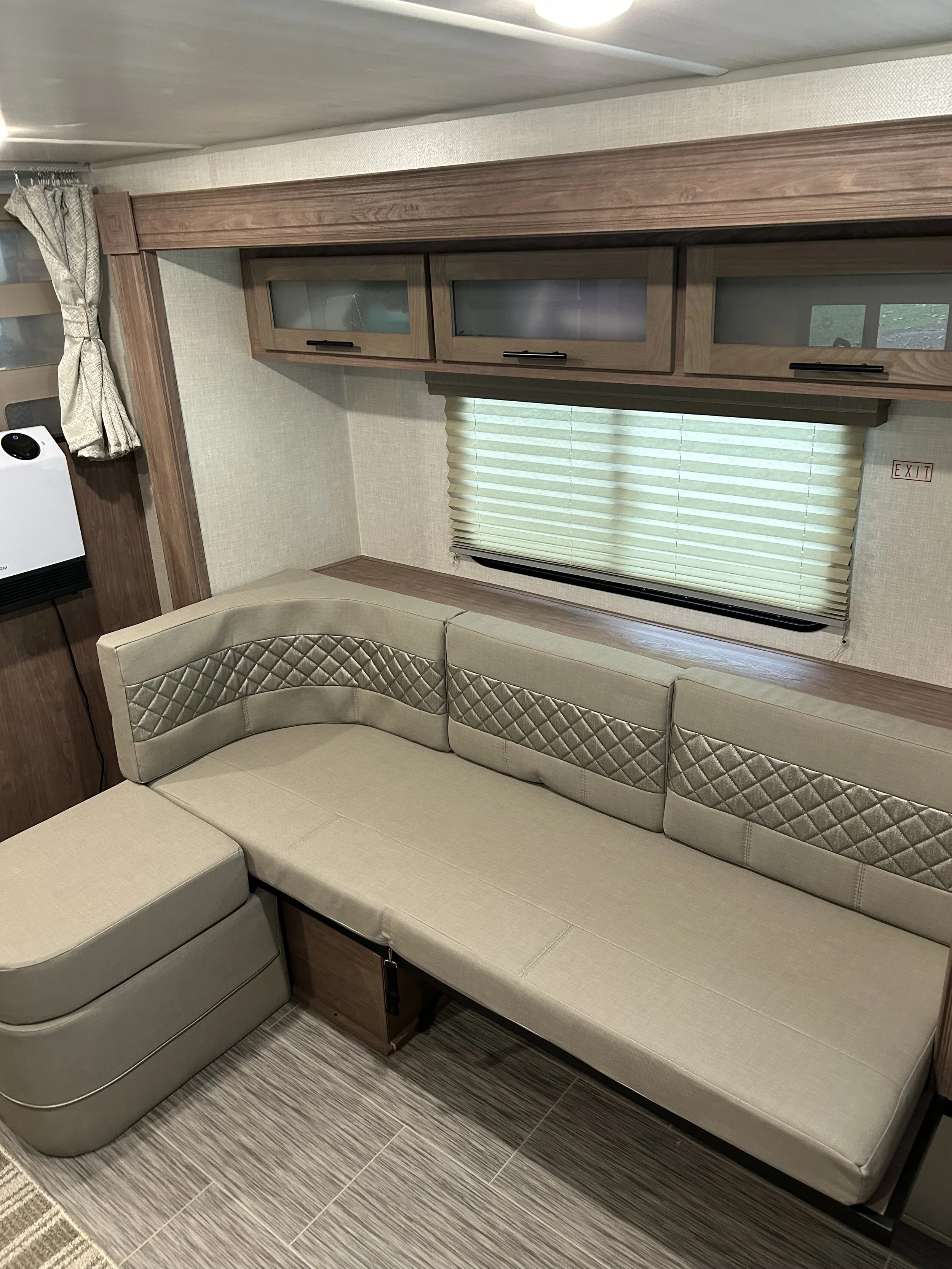 Interior of a recreational vehicle or camper showing a beige upholstered sofa with diamond quilted pattern, a window with blinds, and overhead wooden cabinets with glass inserts.