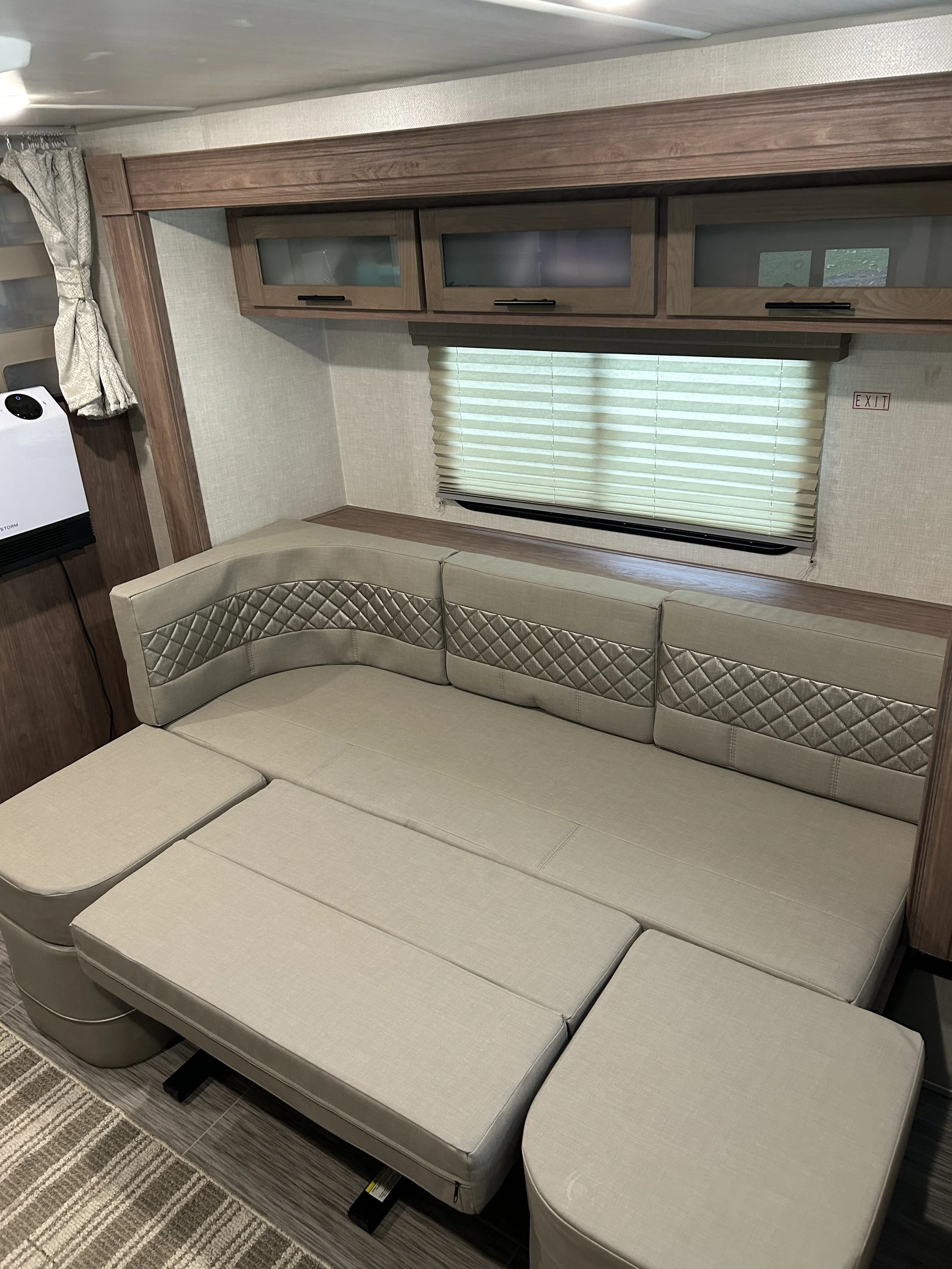 Interior of an RV or camper with beige sectional sofa, upper wooden cabinets, a window with blinds, and a small TV on the side.