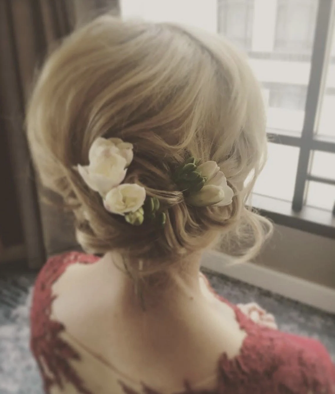 Back view of a woman with blonde hair styled in an elegant updo decorated with white flowers, wearing a reddish dress, sitting near a window.