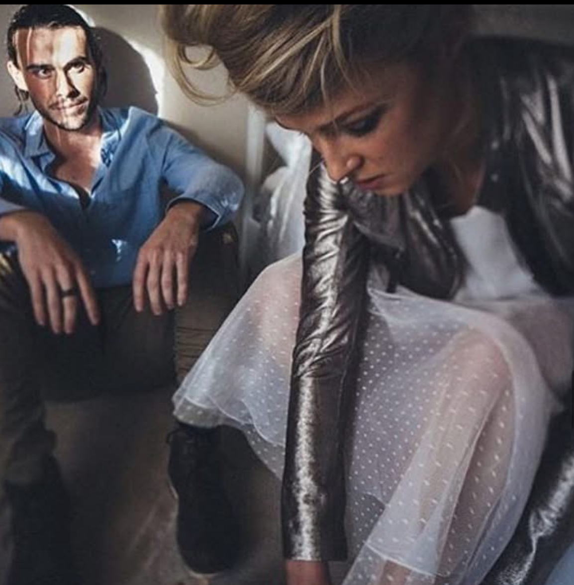 A woman with blonde hair, wearing a shiny silver jacket and a white dress with polka dots, is leaning forward and looking down. A man with dark hair, a beard, and wearing a blue shirt is sitting nearby, resting his arm on his knee, looking at her.