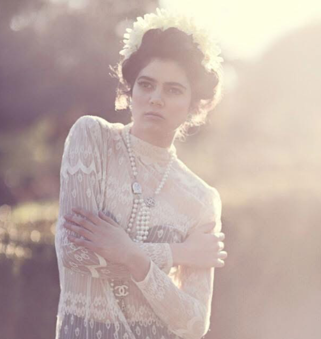 A woman with dark, curly hair wearing a white lace dress, pearl necklaces, and a flower crown, standing outdoors with sunlight behind her.