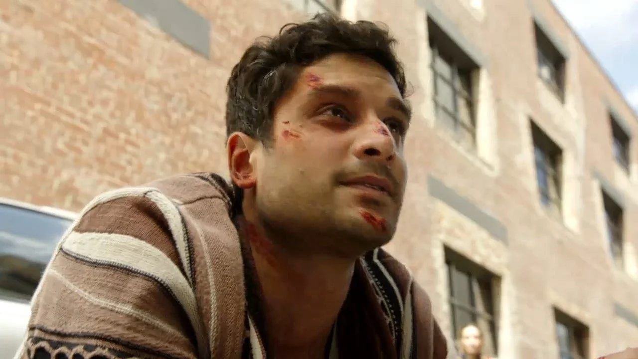 A young man with short dark hair, wearing a brown and beige striped sweater, has scratches and bruises on his face, standing outside in front of a brick building.