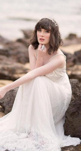 A woman with dark, wavy hair and bangs, wearing a white dress, sitting on rocks by the water with a distant expression.