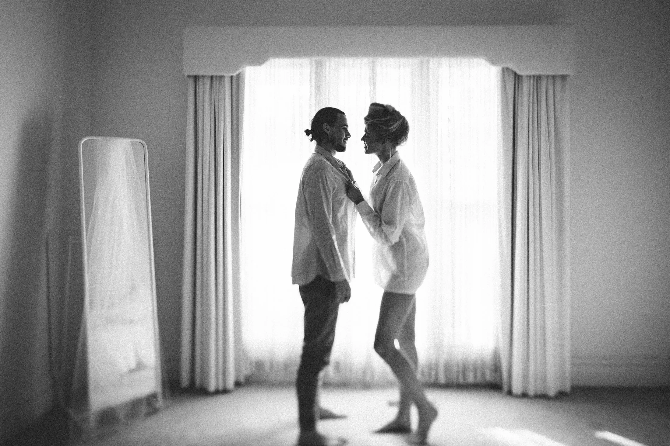 Silhouette of a couple facing each other in a bedroom, with curtains and a mirror in the background, backlit by natural light.