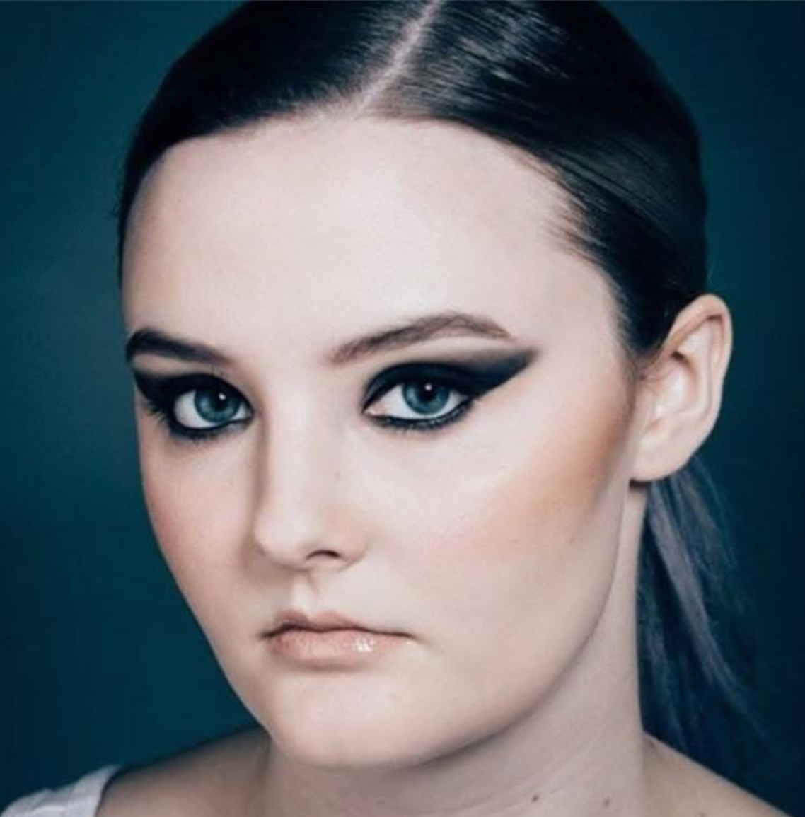A woman with dark hair and striking makeup, including bold black eyeliner and eyeshadow, looking directly at the camera.