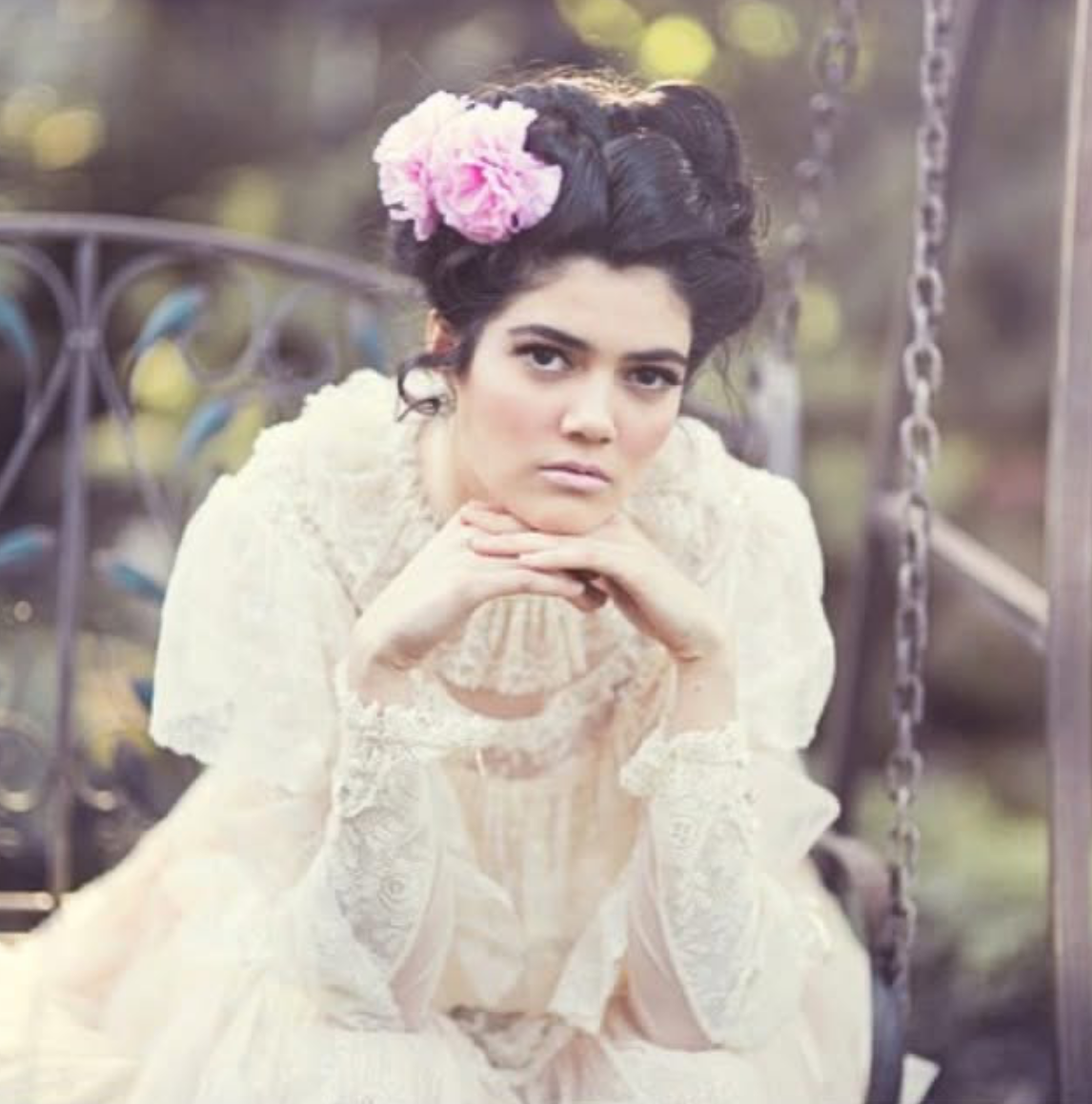 A woman in a vintage lace dress with dark hair styled in an updo, adorned with pink flowers, sits on a swing with chains in an outdoor setting.