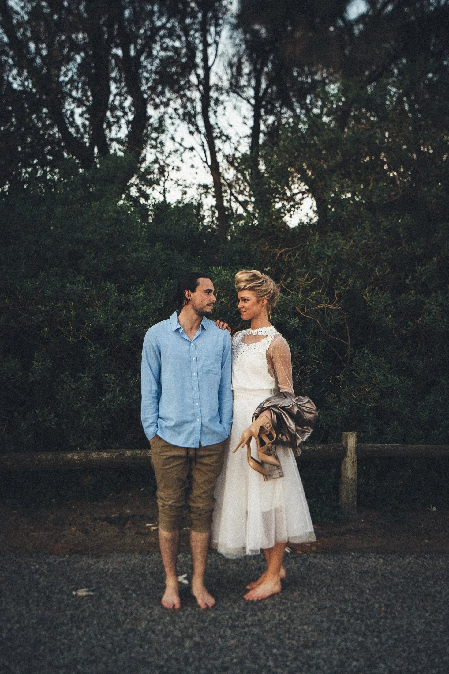 A man and a woman standing barefoot on a paved road, with a lush green forest in the background. The woman is wearing a white lace dress and holding a pair of high-heeled shoes, while the man is dressed casually in a blue shirt and khaki shorts. They