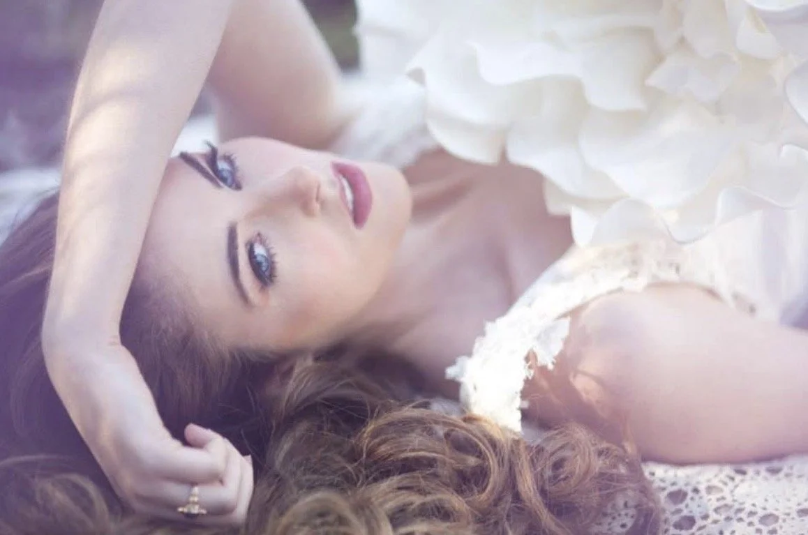 A woman with long, wavy hair lying on her back, wearing a white dress with floral details, looking at the camera.