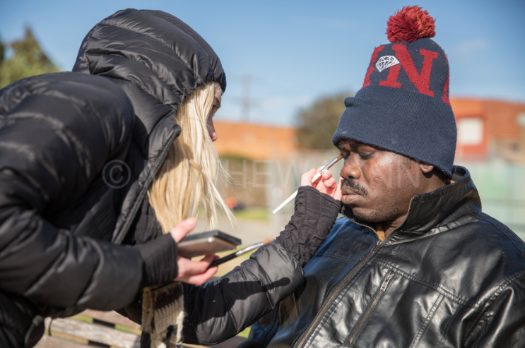 A woman applying makeup or face paint to a man outdoors on a sunny day, with a blue sky and some trees and buildings in the background. Both are dressed warmly, and the man is wearing a knit winter hat with a pom-pom and a leather jacket.