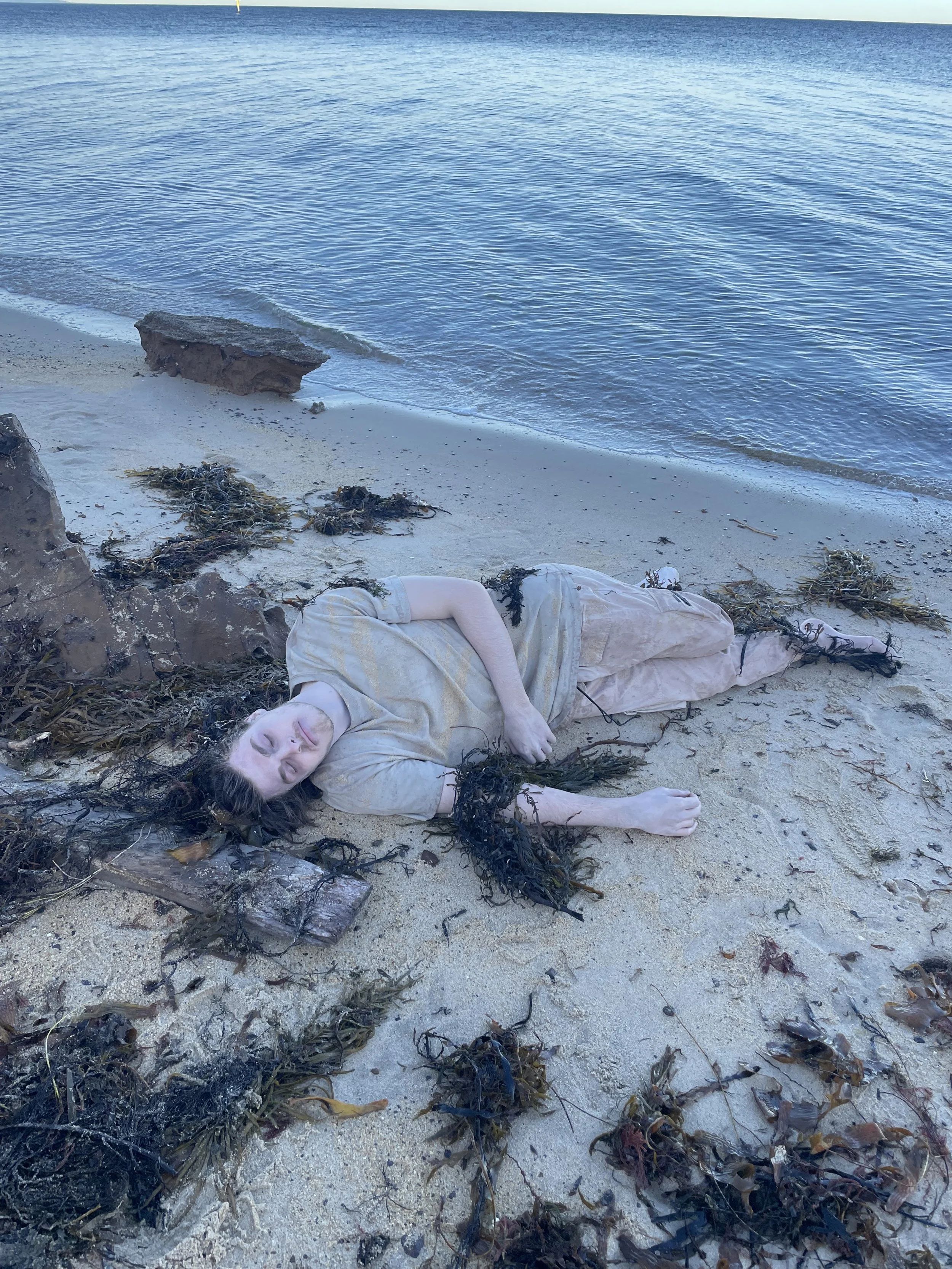 Person lying on the sandy beach surrounded by seaweed and rocks, near the water's edge.