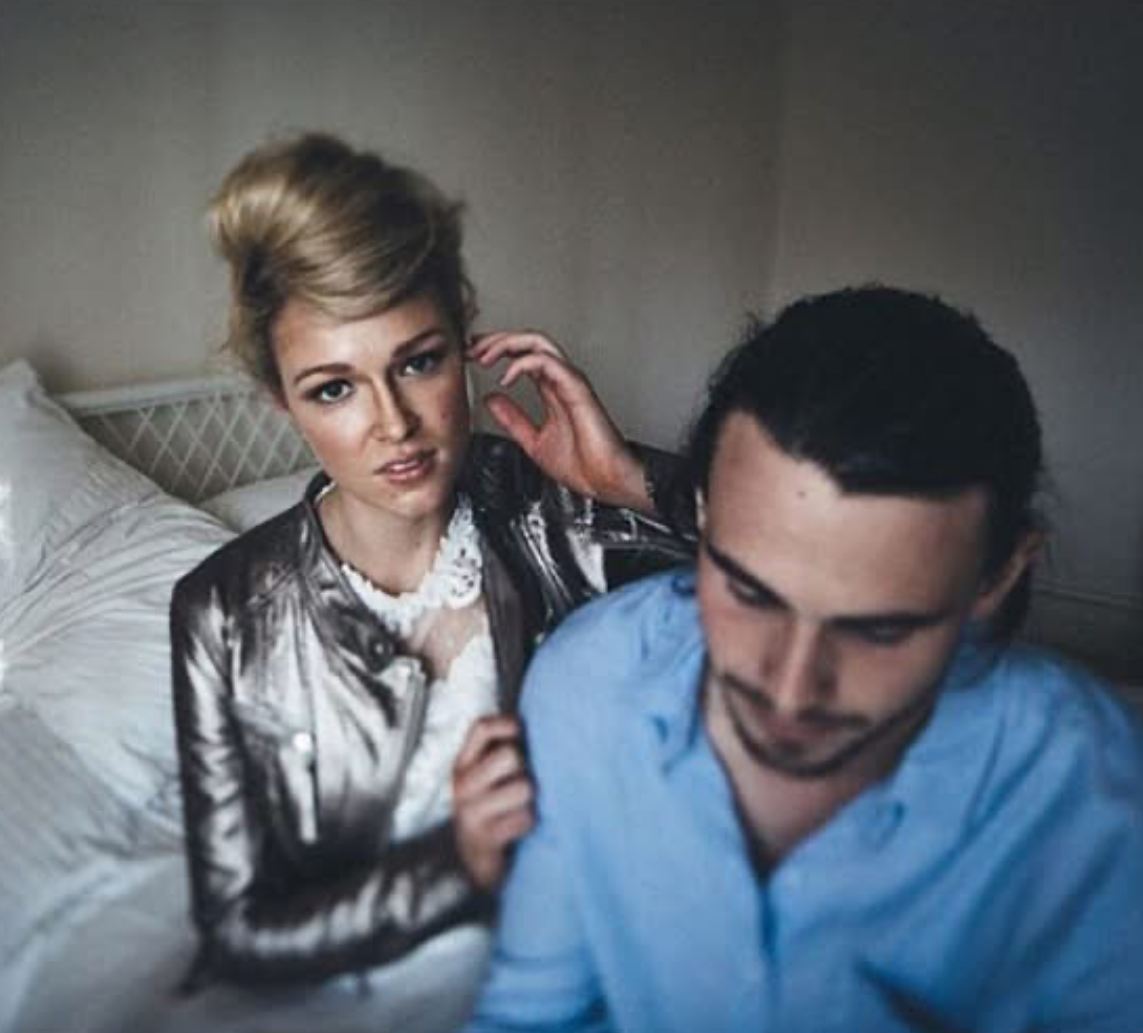 A woman with blonde hair styled in an updo, wearing a shiny jacket and white lace top, is sitting beside a man with dark hair in a blue shirt. The woman is touching her hair while looking at the camera, and the man is looking down, seated on a bed in