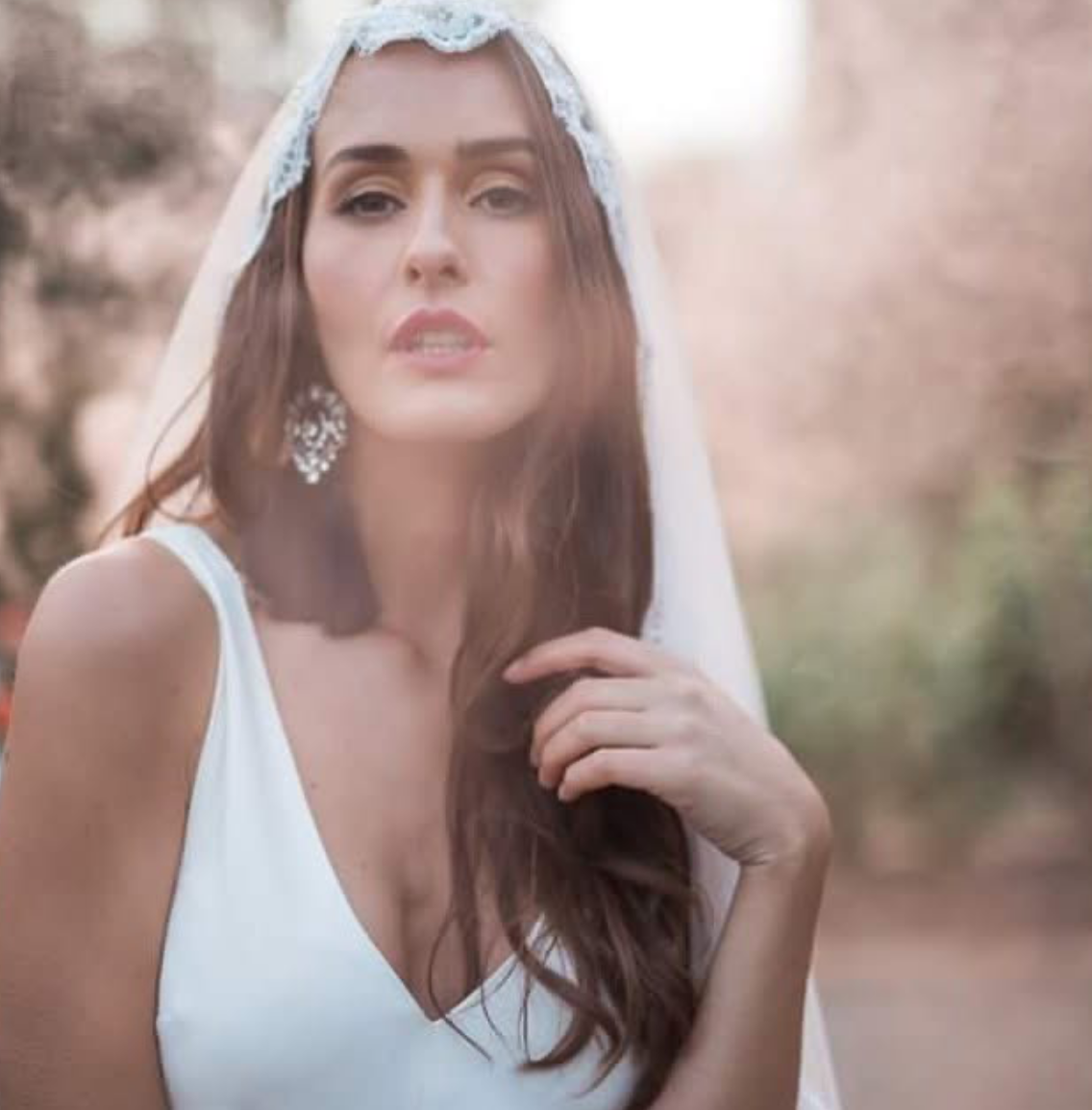 Portrait of a woman in a white dress with a lace veil, outdoors with trees in the background.