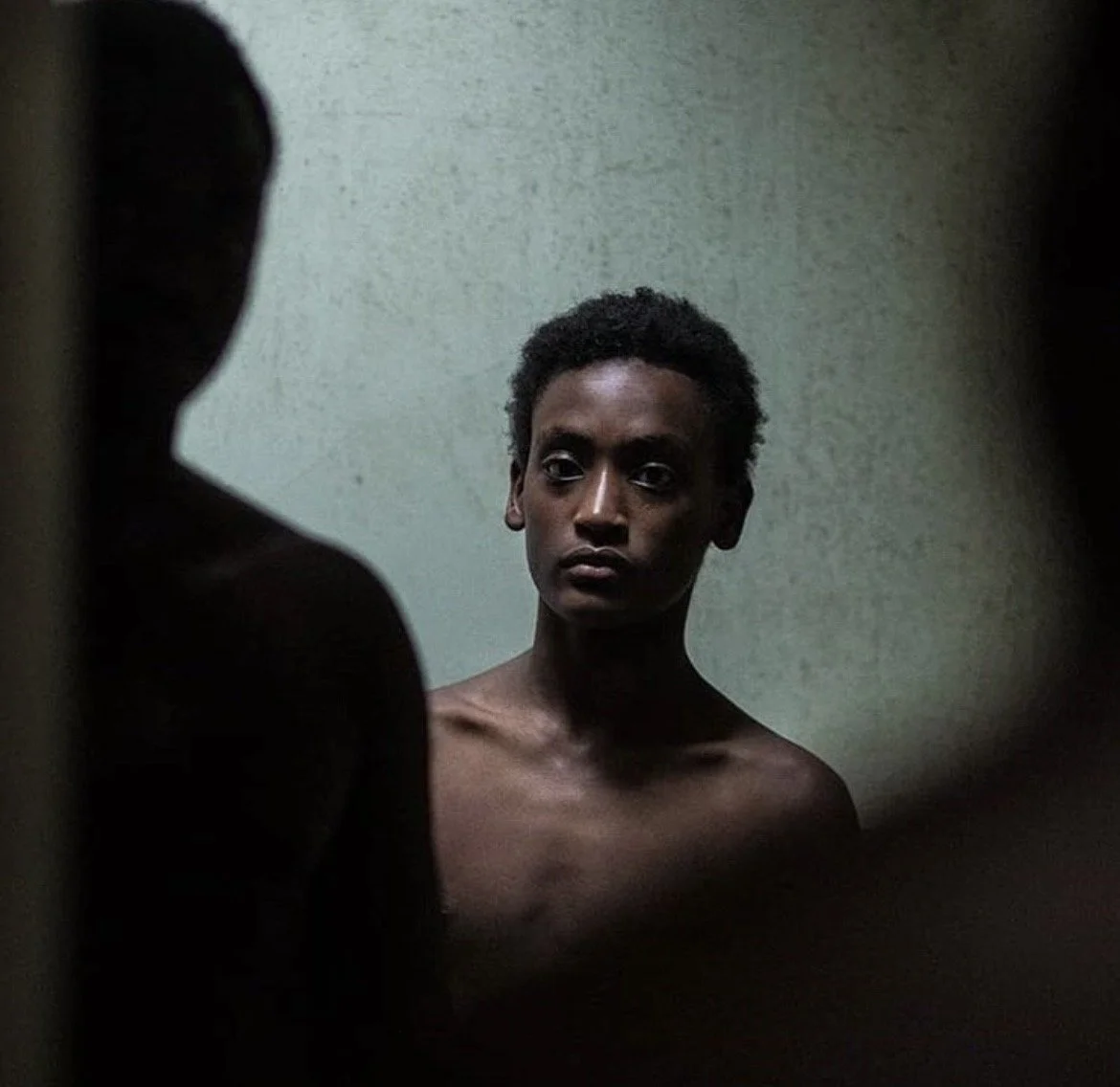 A young man with short curly hair looking intently at his reflection in a mirror, with a plain wall in the background.