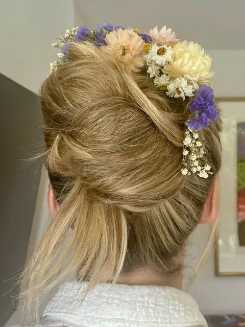 Woman with blonde hair styled in an elegant updo adorned with a floral crown of white, cream, purple, and blue flowers.