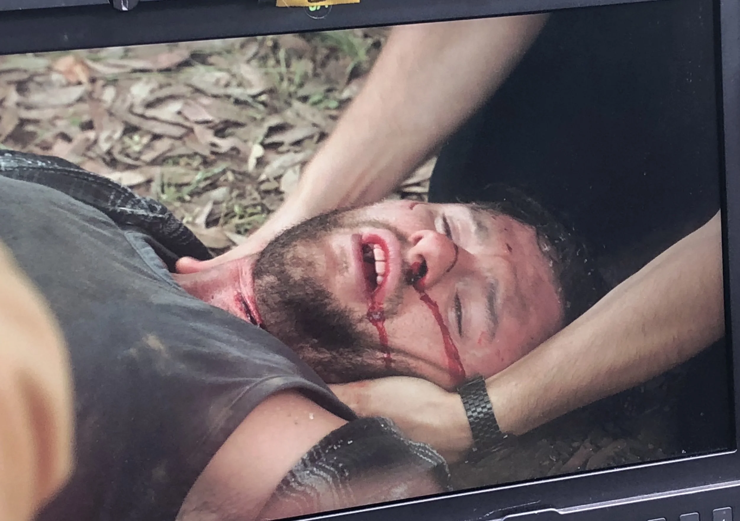 A man with a beard lying on the ground with blood streaming from his nose, eyes closed, with someone holding his head and another person holding his arm, on a forest floor covered with leaves.