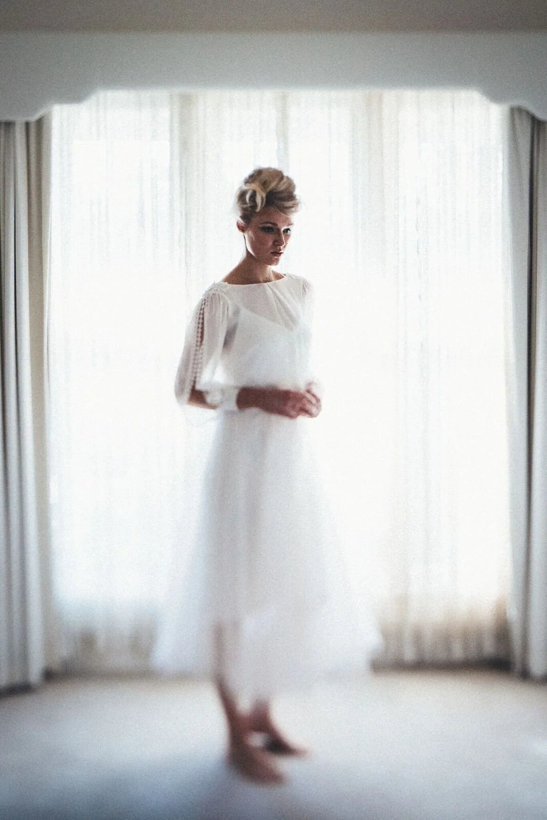 A woman in a white dress standing indoors near a window with sheer curtains, viewed through a mirror.