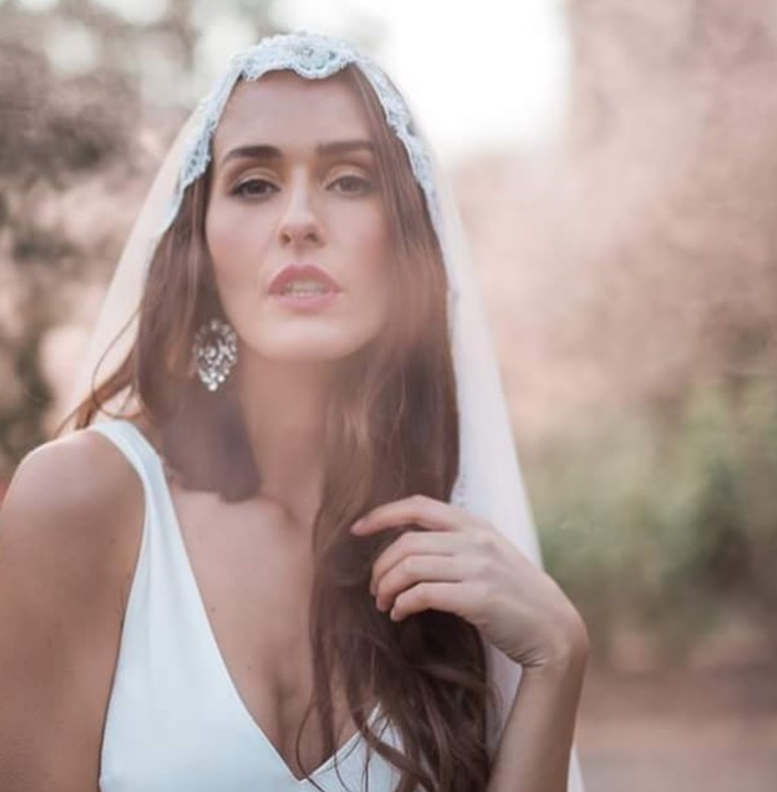 A woman wearing a white dress and a lace veil, standing outdoors with blurred foliage in the background, looking directly at the camera.