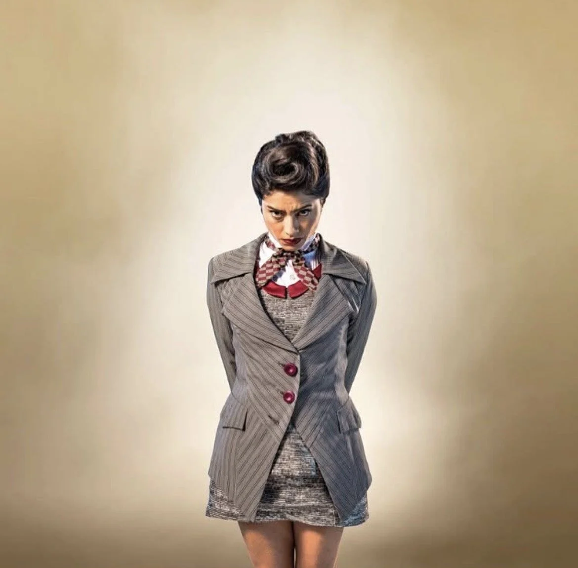 Woman with dark hair styled in a vintage 1950s updo, wearing a classic pinstripe suit jacket, a patterned skirt, white blouse with a bow tie, and red lipstick, standing with a stern expression against a plain beige background.