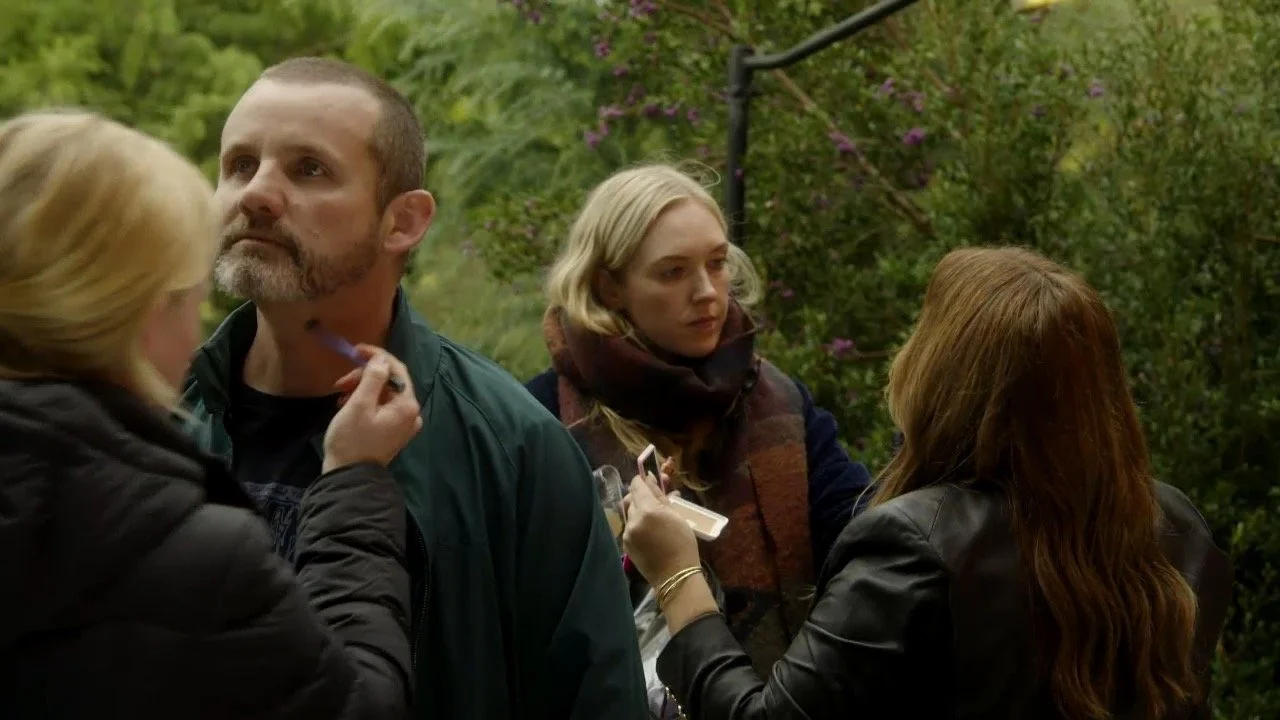 Four people, two women and two men, outdoors in a green, leafy area, engaged in conversation and makeup application.