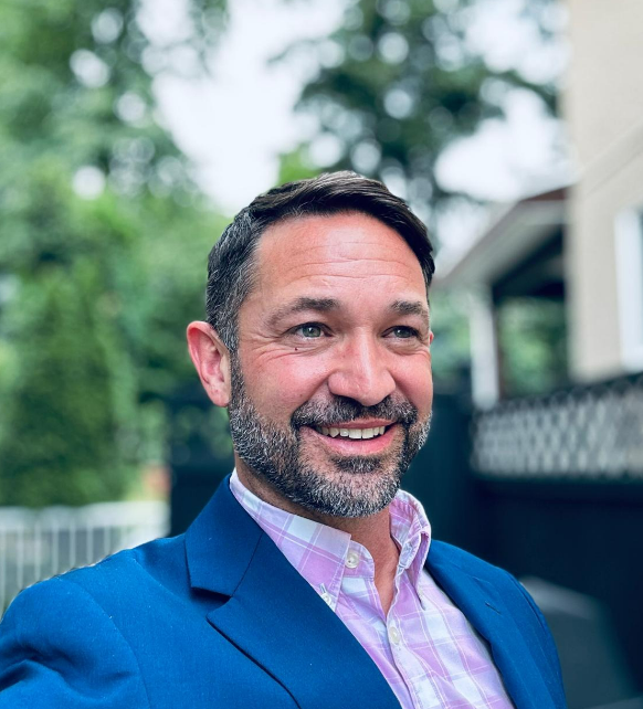 A smiling man in a pink checkered shirt and blue blazer outdoors, with trees and a house in the background.