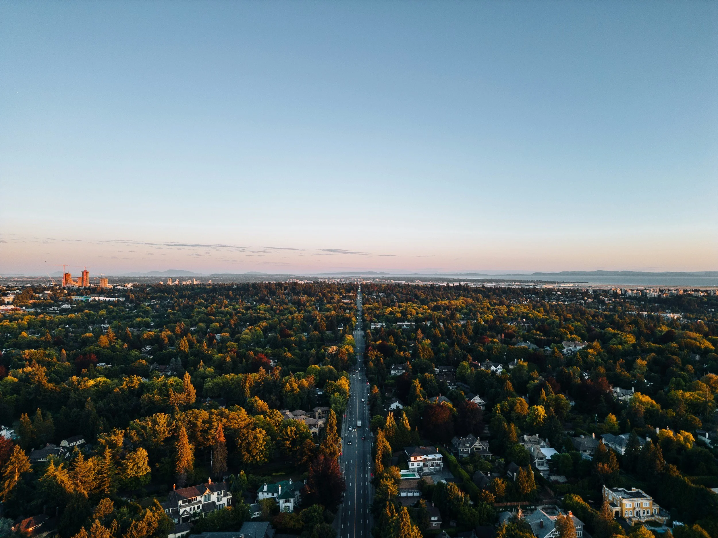 An aerial view of a city with a long straight road running through a mostly green residential area, with taller buildings in the distance, under a clear sky at sunset.