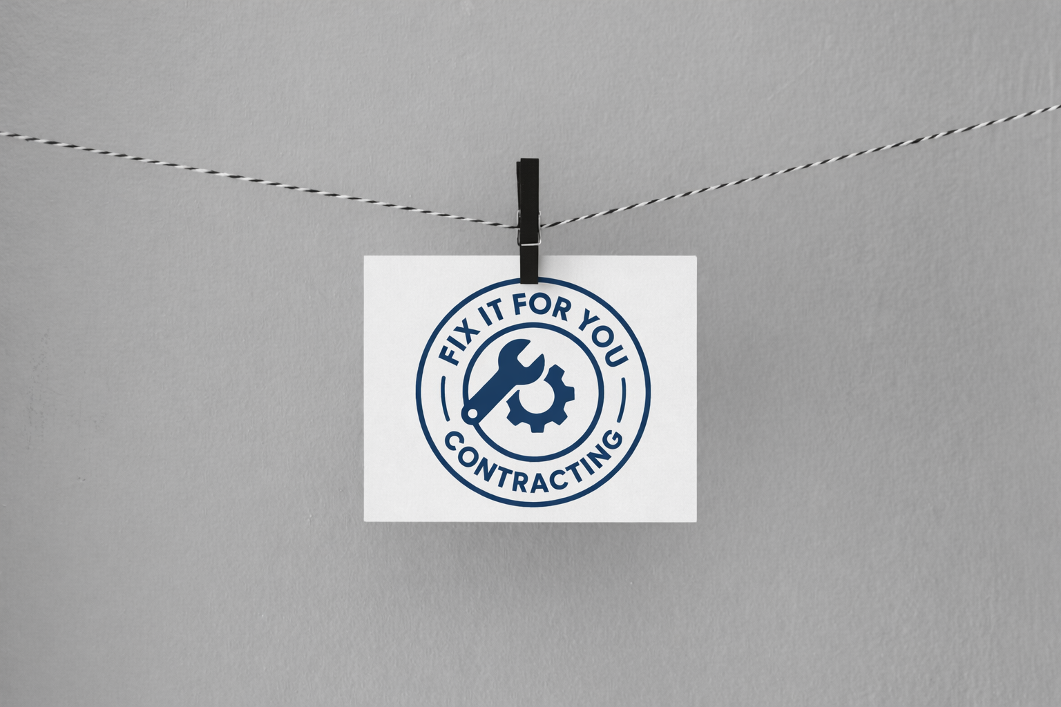 A white business card with a blue circular logo hangs from a black clothespin on a string. The logo contains a wrench and gear icon with the text 'Fix It For You Contracting.'