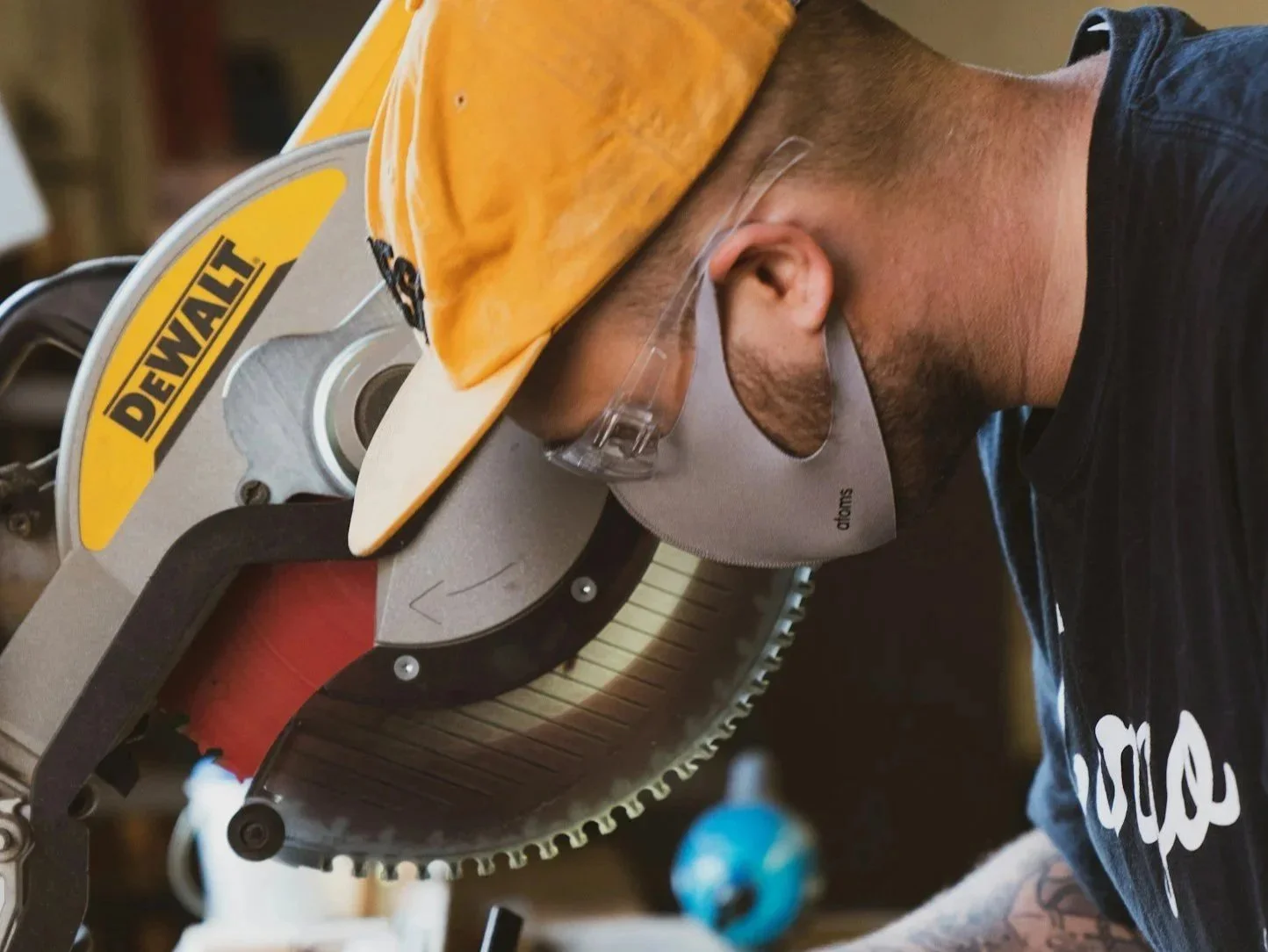 A person wearing a yellow safety helmet and glasses is cutting wood with a circular saw in a workshop.
