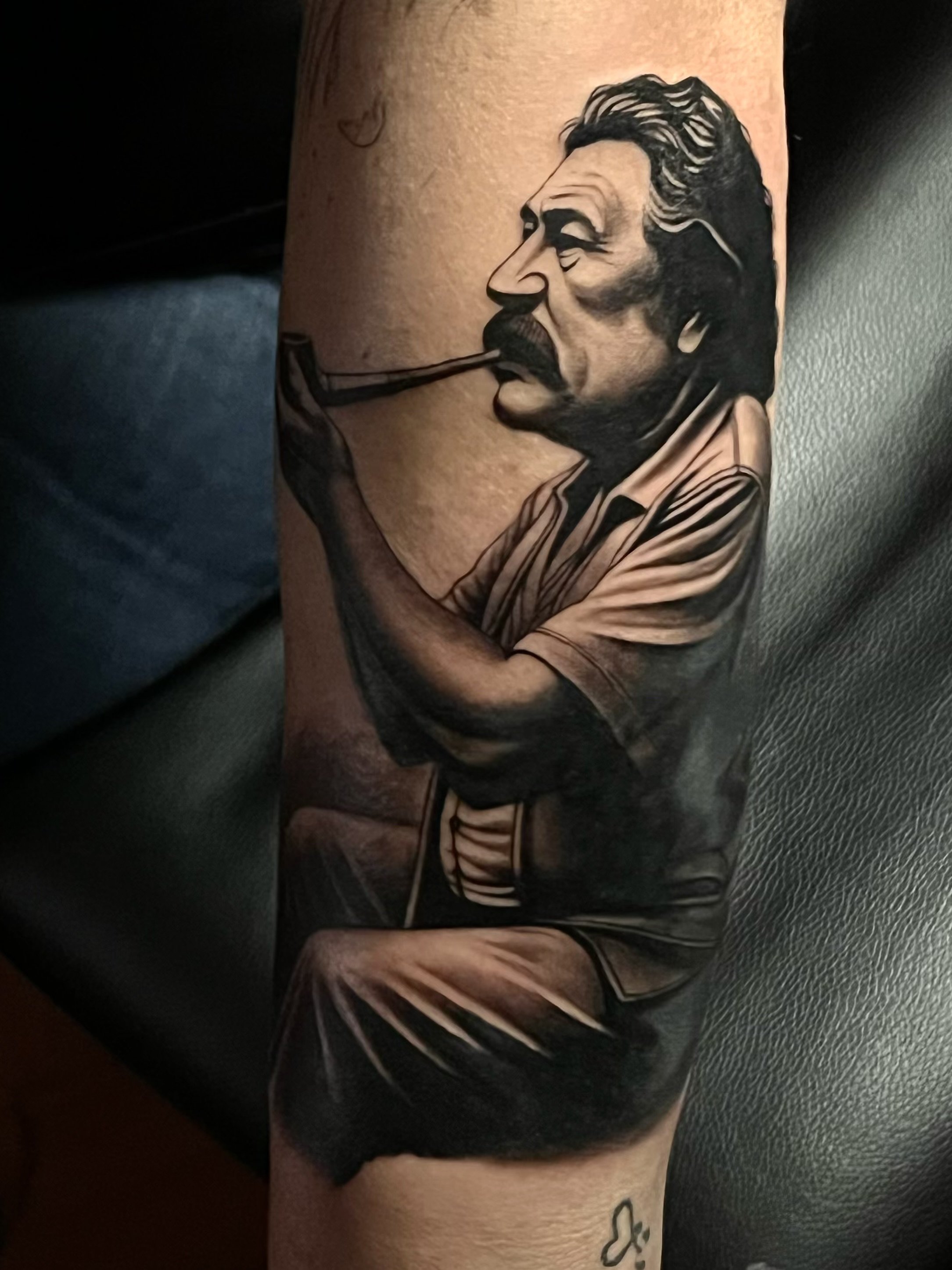 Tattoo of a man with wavy hair and a mustache, smoking a pipe, on a person's arm.