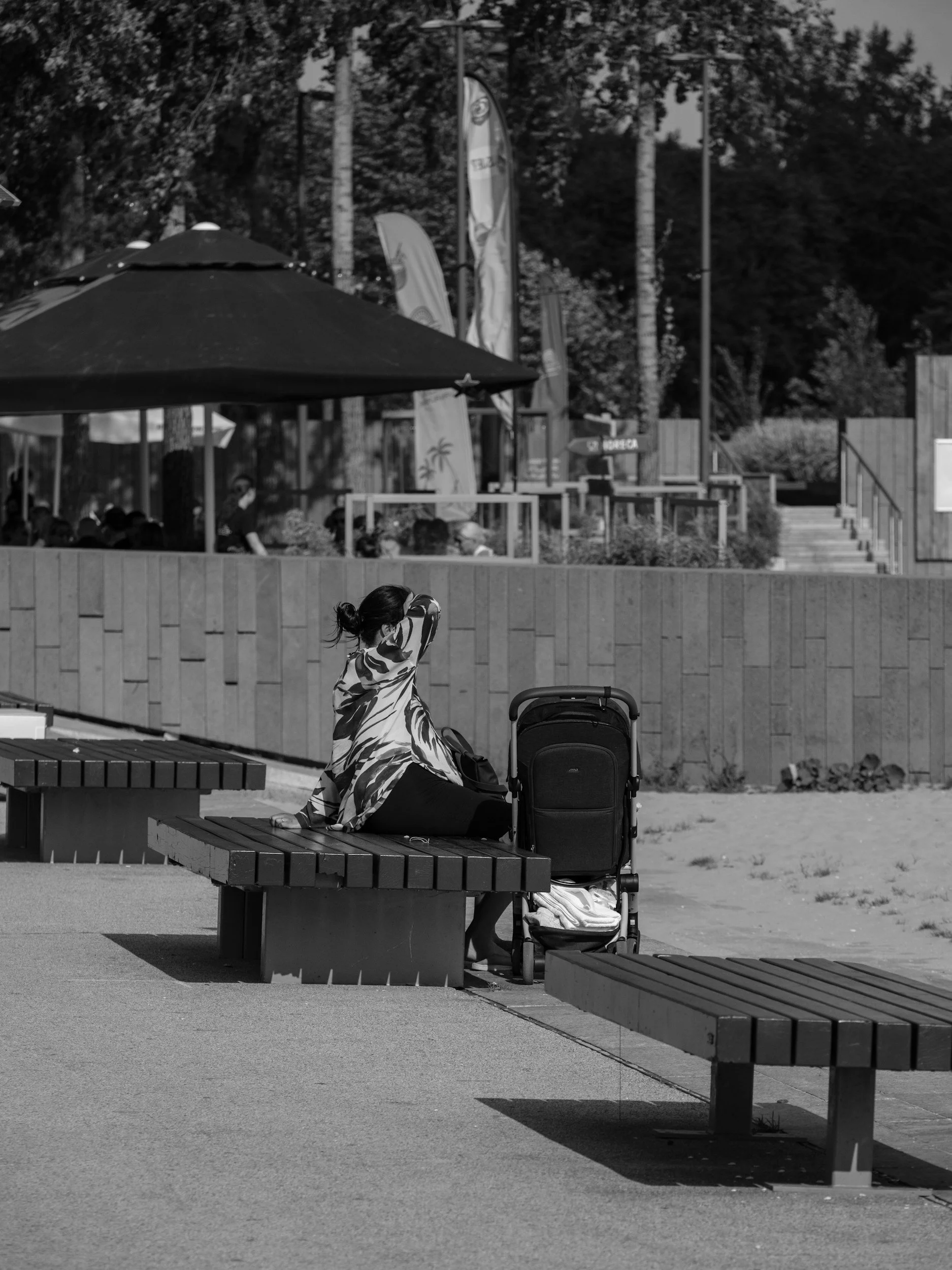 A woman sitting on a bench with a baby stroller beside her in an outdoor park setting. She is wearing a patterned shawl or scarf.