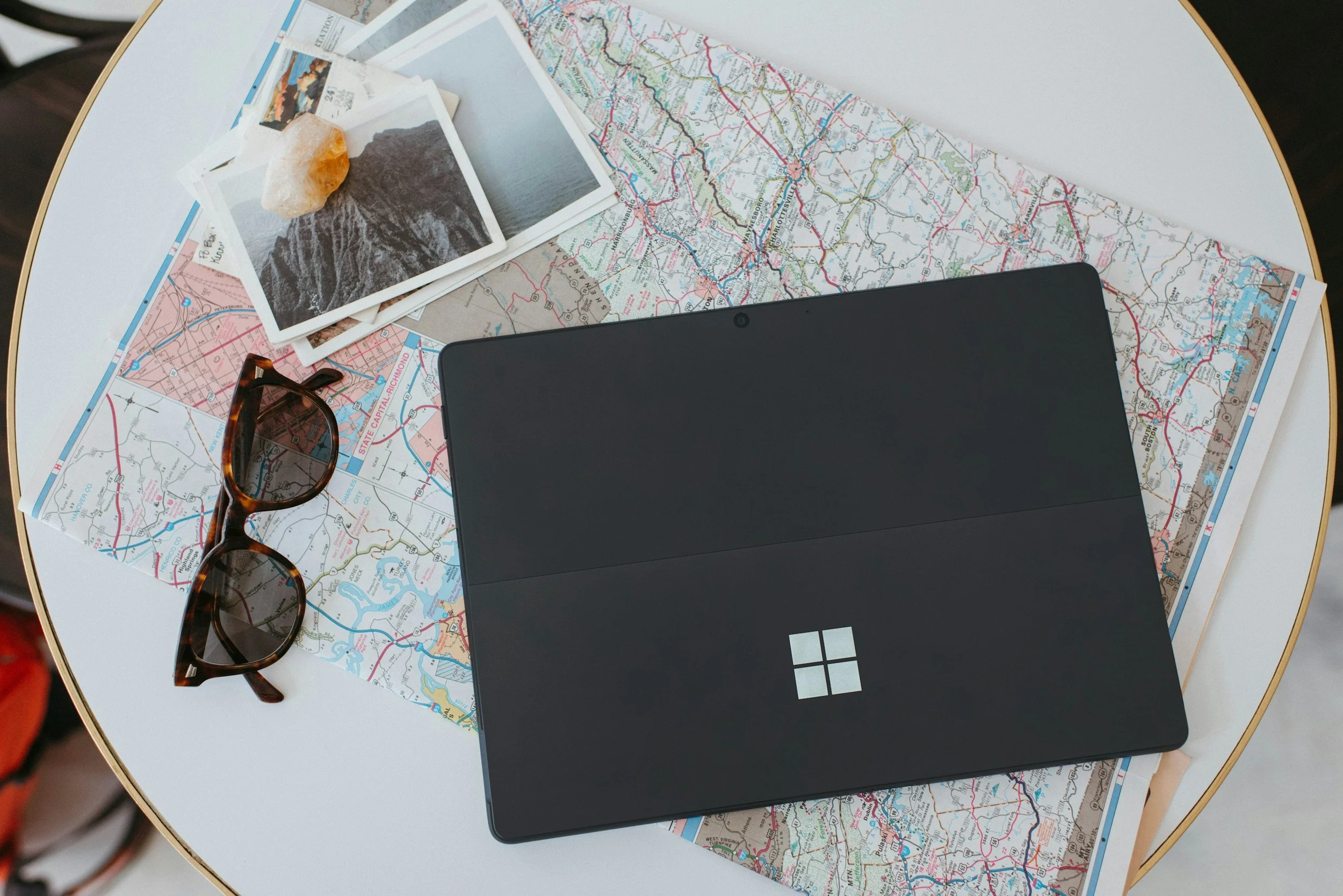 A laptop with a Microsoft logo on the lid is placed on top of printed maps, with a pair of sunglasses and small travel photographs nearby. An orange rock sits on one of the photographs.