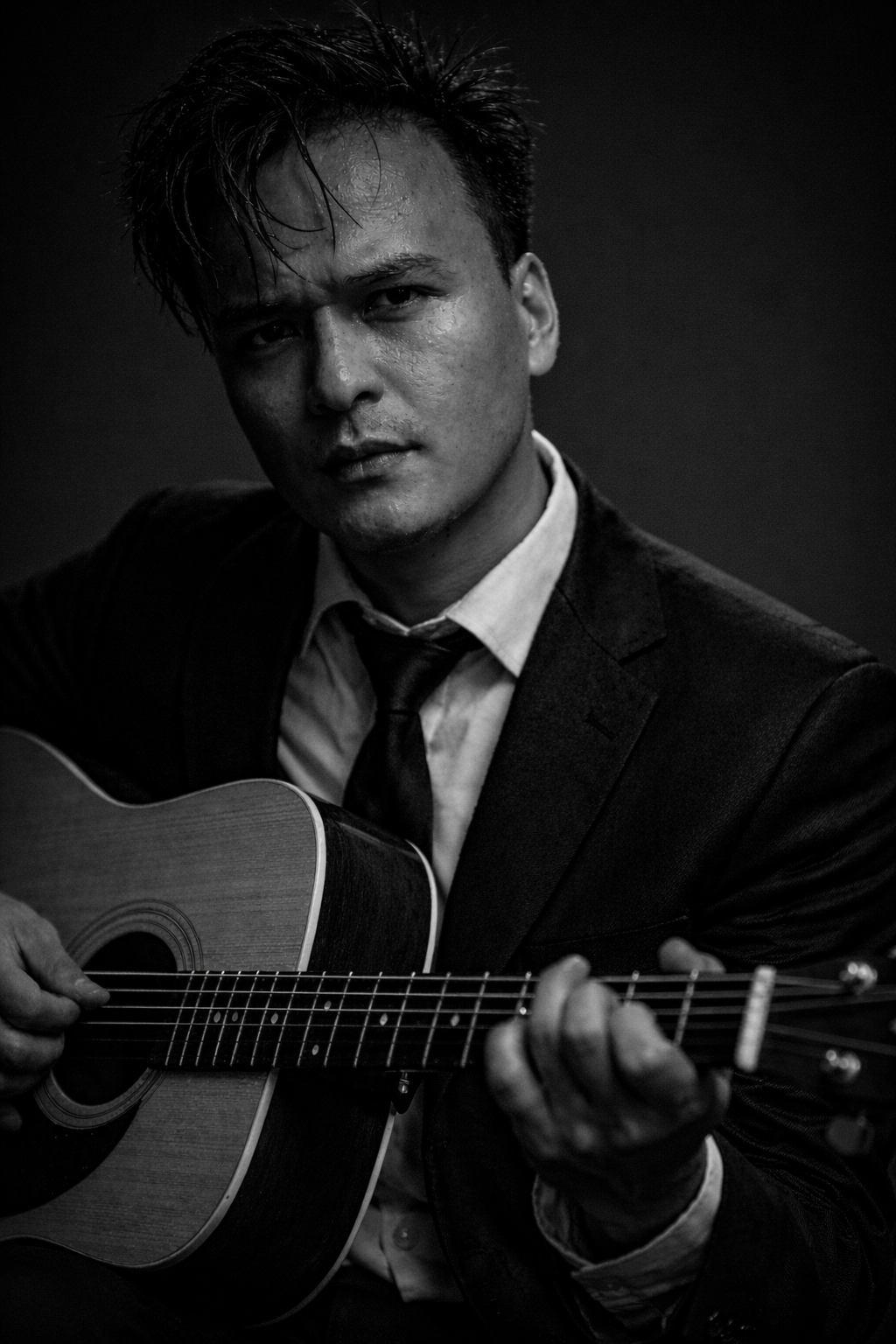A black and white photo of a man in a suit playing an acoustic guitar, with a serious expression on his face.