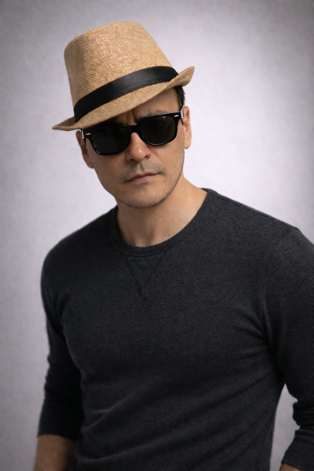 A man wearing a straw fedora hat with a black band, black sunglasses, and a dark long-sleeve shirt, standing against a plain, light-colored background.