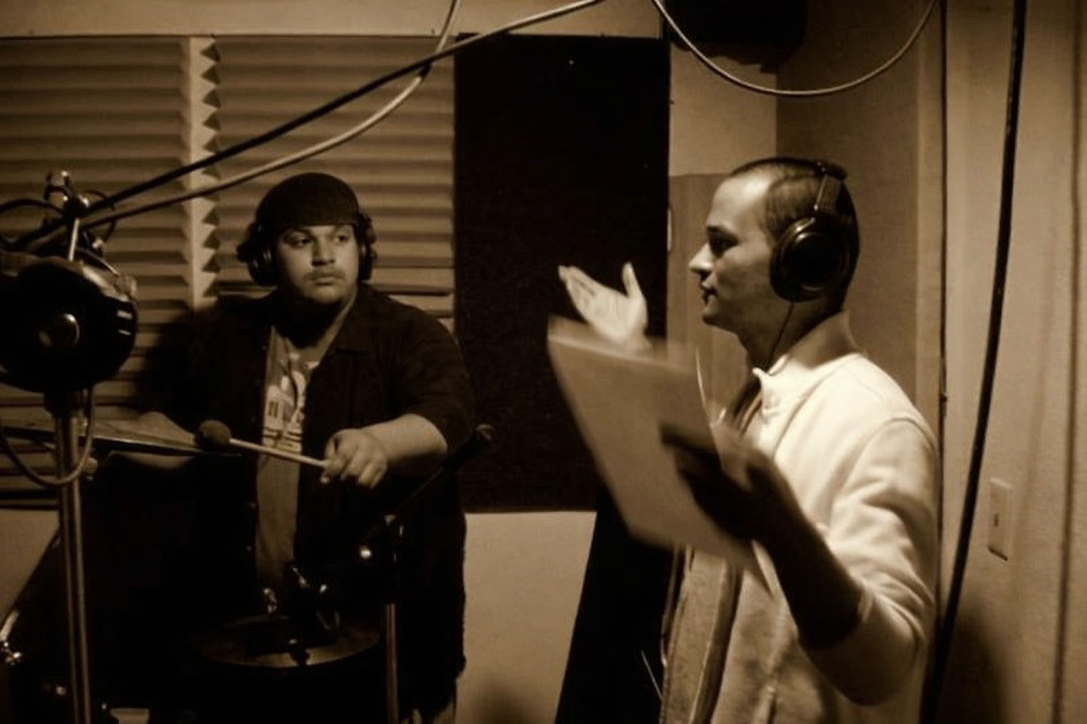 Two men in a recording studio, one is holding a script and talking, the other is listening with headphones.