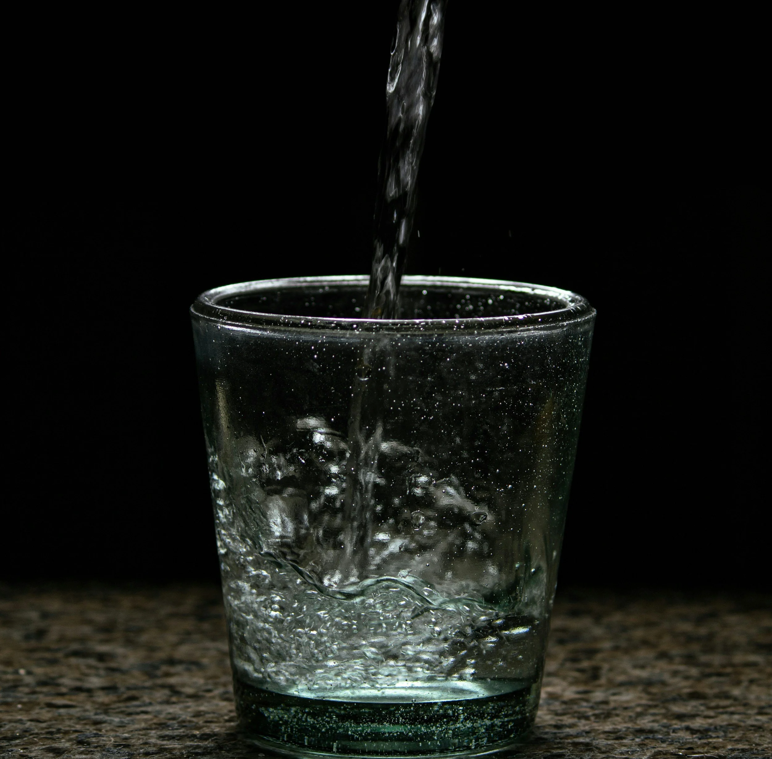 A glass being filled with water from a pouring stream against a black background.