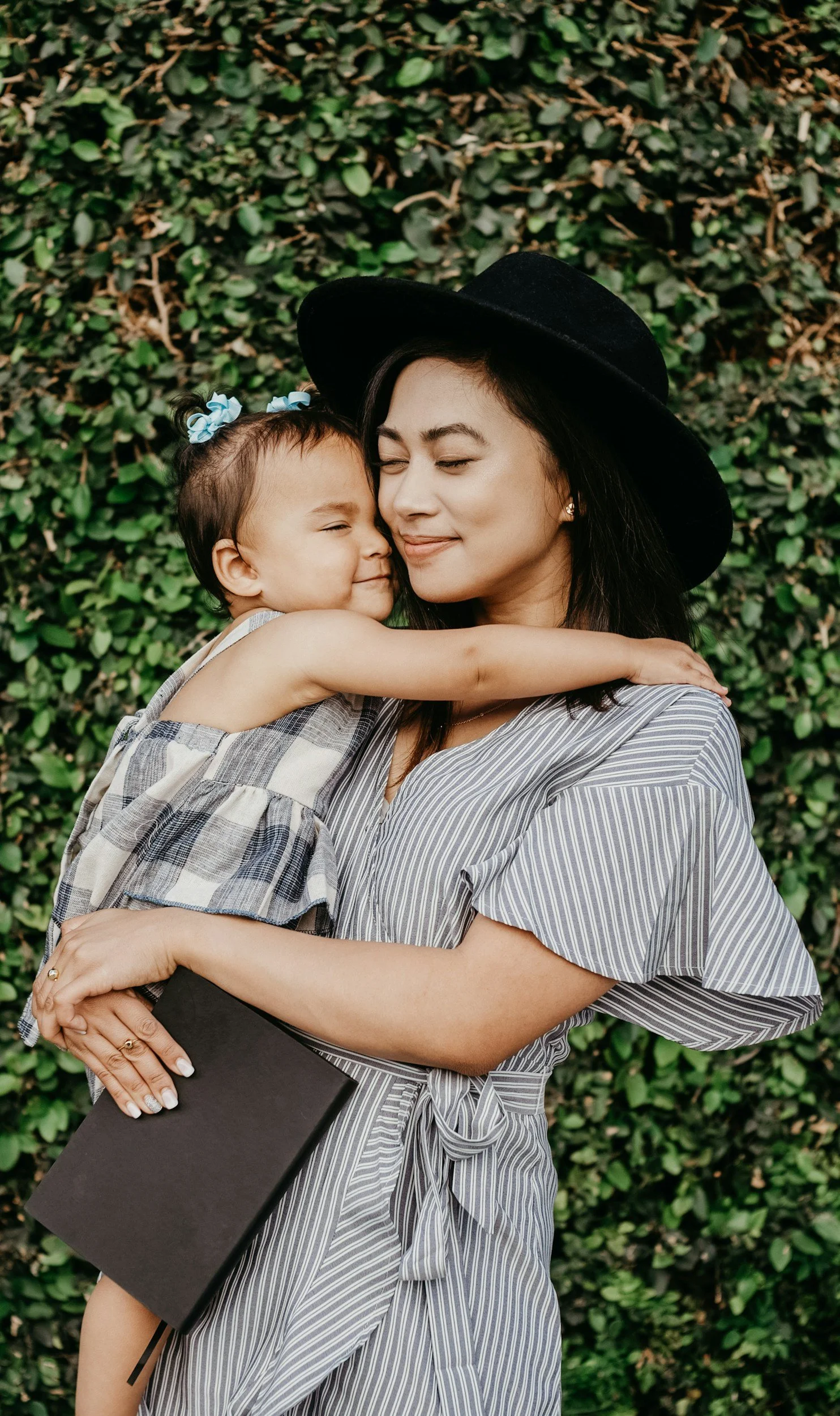 A woman holding a young girl in front of a leafy green hedge, both with eyes closed and smiling, sharing a hug.
