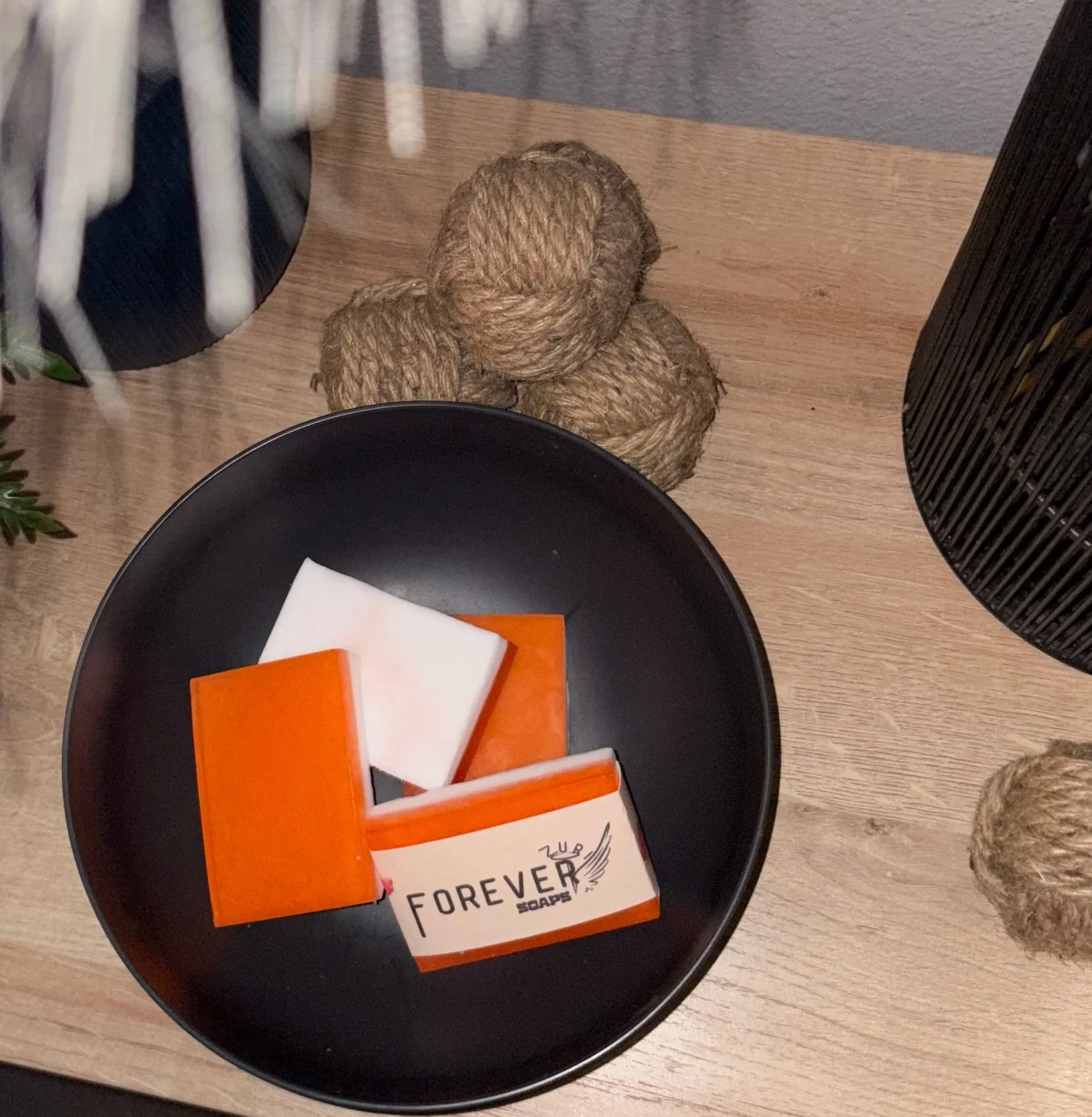 A black bowl with orange, white, and beige soap bars on a wooden surface, with twine balls and part of a black wire basket nearby.