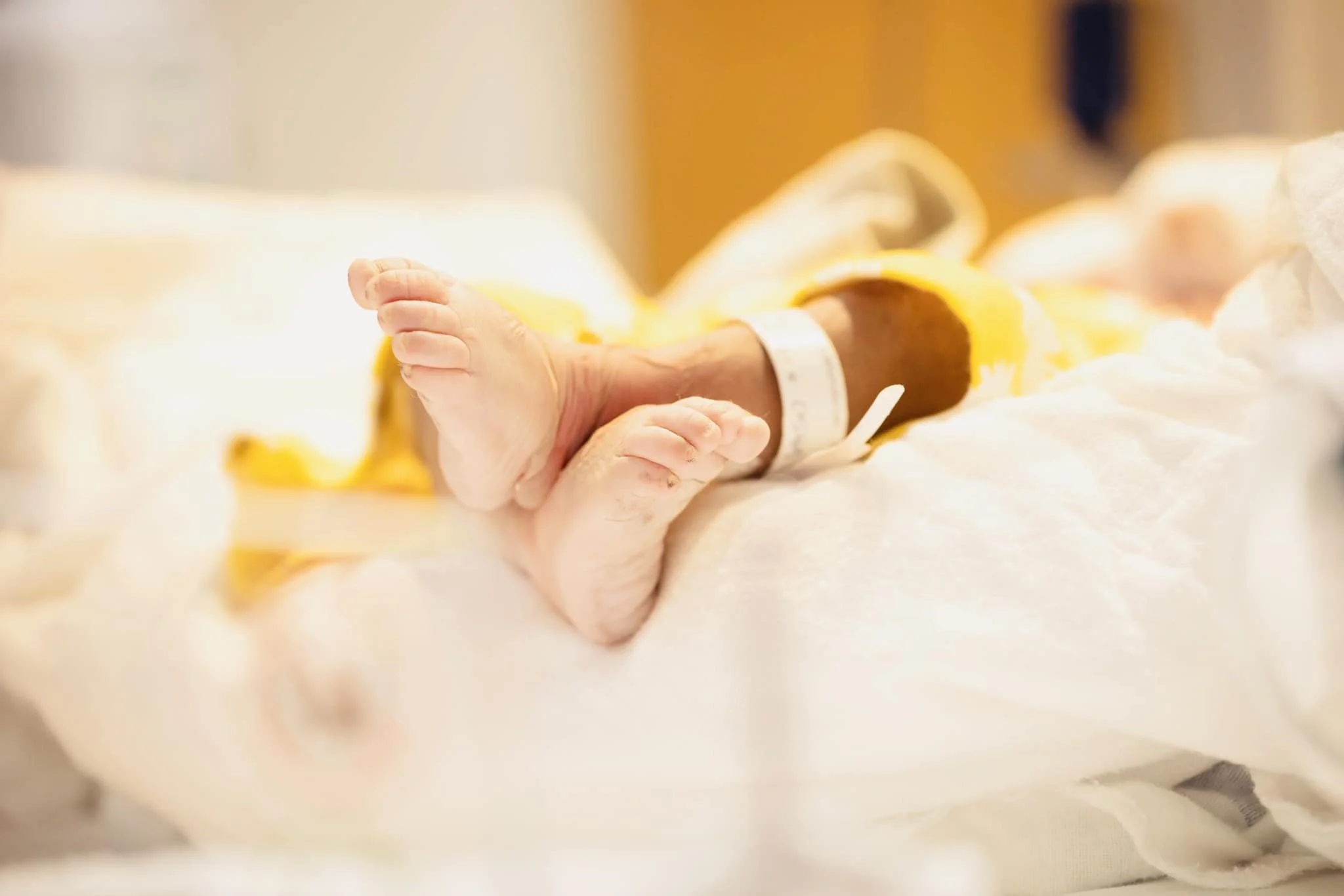 Close-up of a newborn baby's feet with hospital identification bands, lying on a hospital bed or incubator.