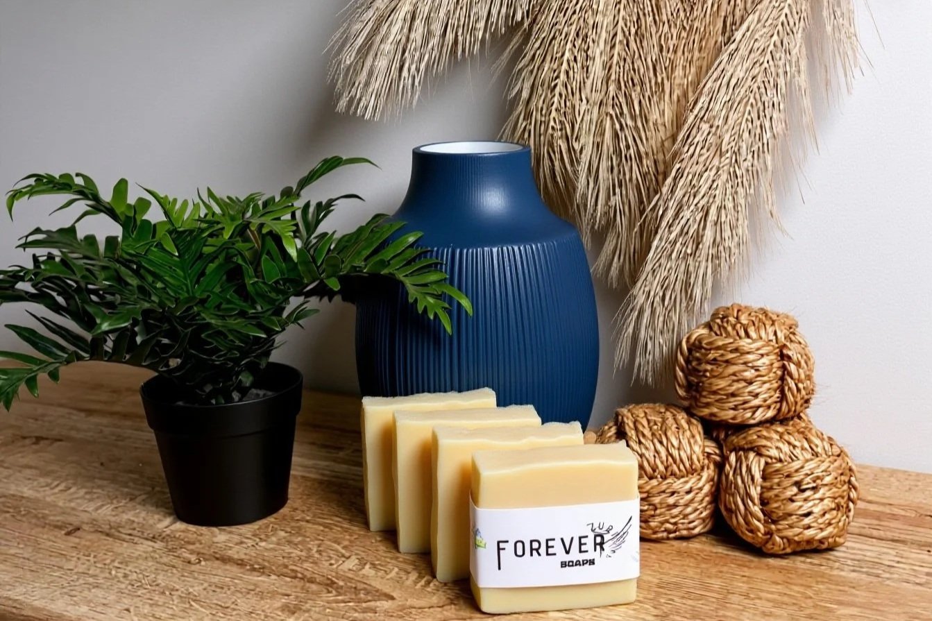 Decorative arrangement on wooden surface featuring a potted green plant, a blue vase, natural fiber balls, dried pampas grass, and bar soap labeled 'Forever'.