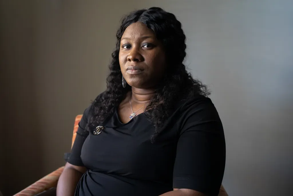 An African American woman with curly black hair wearing a black top, jewelry including earrings, a necklace, and a pin, sitting on a chair against a plain background.
