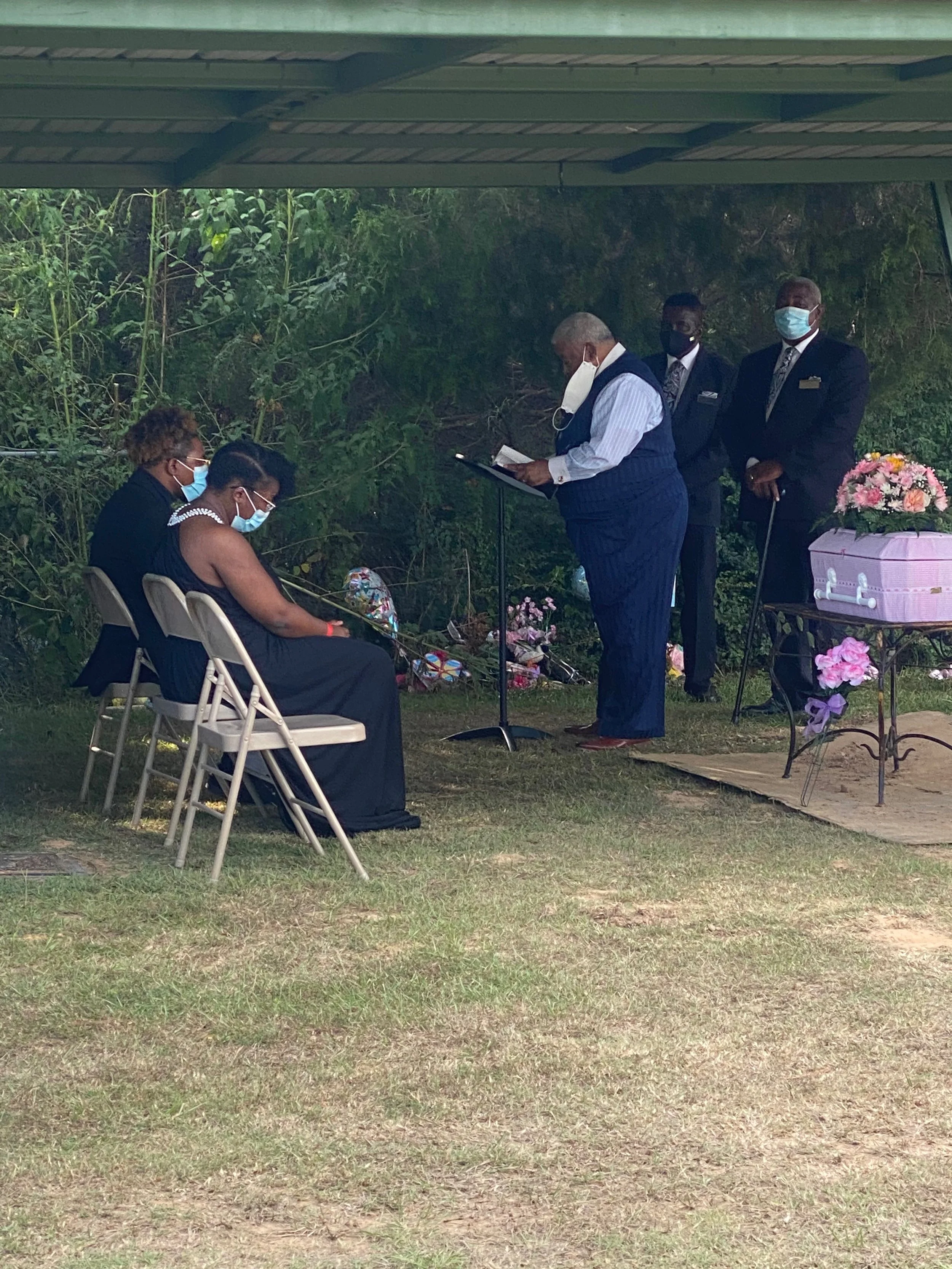 An outdoor funeral scene with five people wearing face masks. Two women are seated on white chairs, one with short curly hair and the other with braided hair. They are dressed in dark clothing, with flowers beside them. Three men are standing; one is reading a paper, and the other two are wearing suits and standing with solemn expressions. There are flowers and a pink casket with floral arrangements on a stand nearby. The scene takes place under a covered area next to trees and greenery.