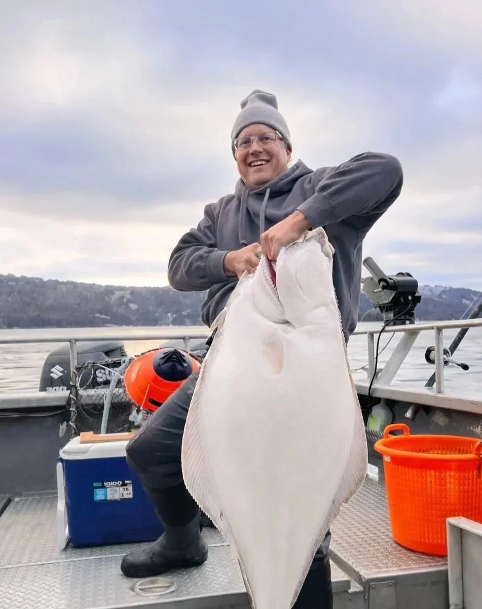 Man on a boat holding a large flat fish, smiling, wearing a gray hoodie, gray beanie, glasses, and rubber boots, with cloudy sky and water in the background.