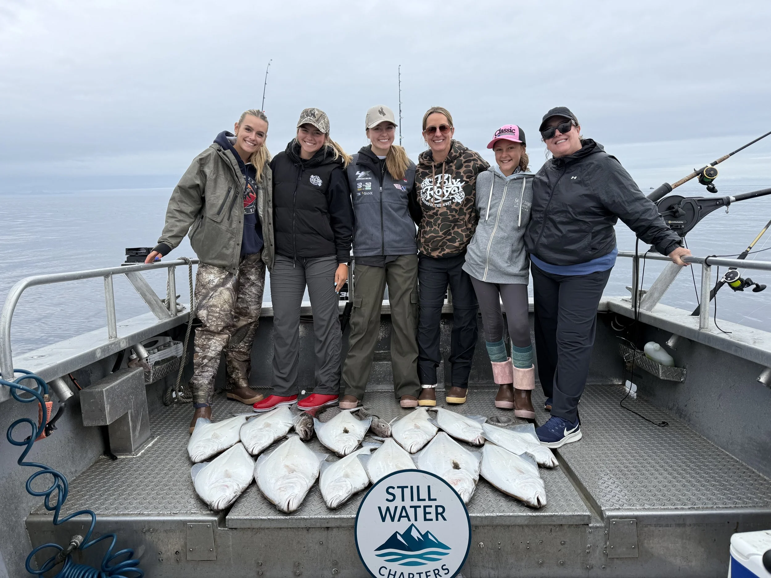 Group of six women on a fishing boat holding freshly caught fish, with fishing rods available, in open water under a cloudy sky, with a logo reading "Still Water Charters" on the boat's floor.