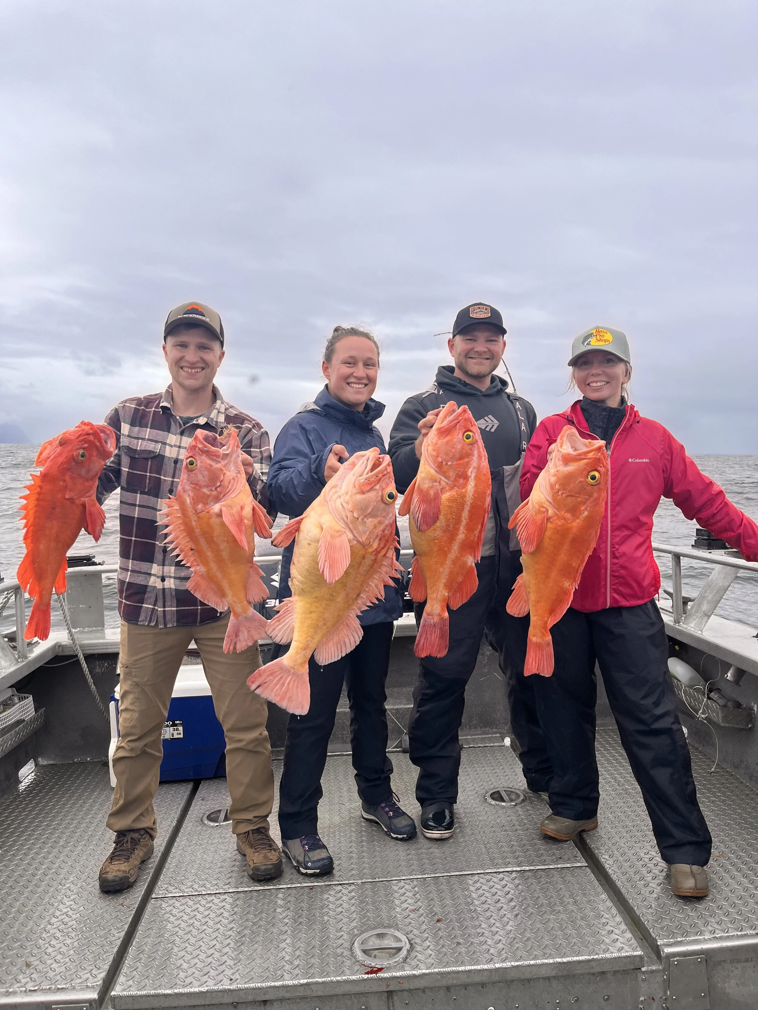 Four people on a boat holding up large orange fish they caught during a fishing trip, with cloudy sky and ocean in the background.