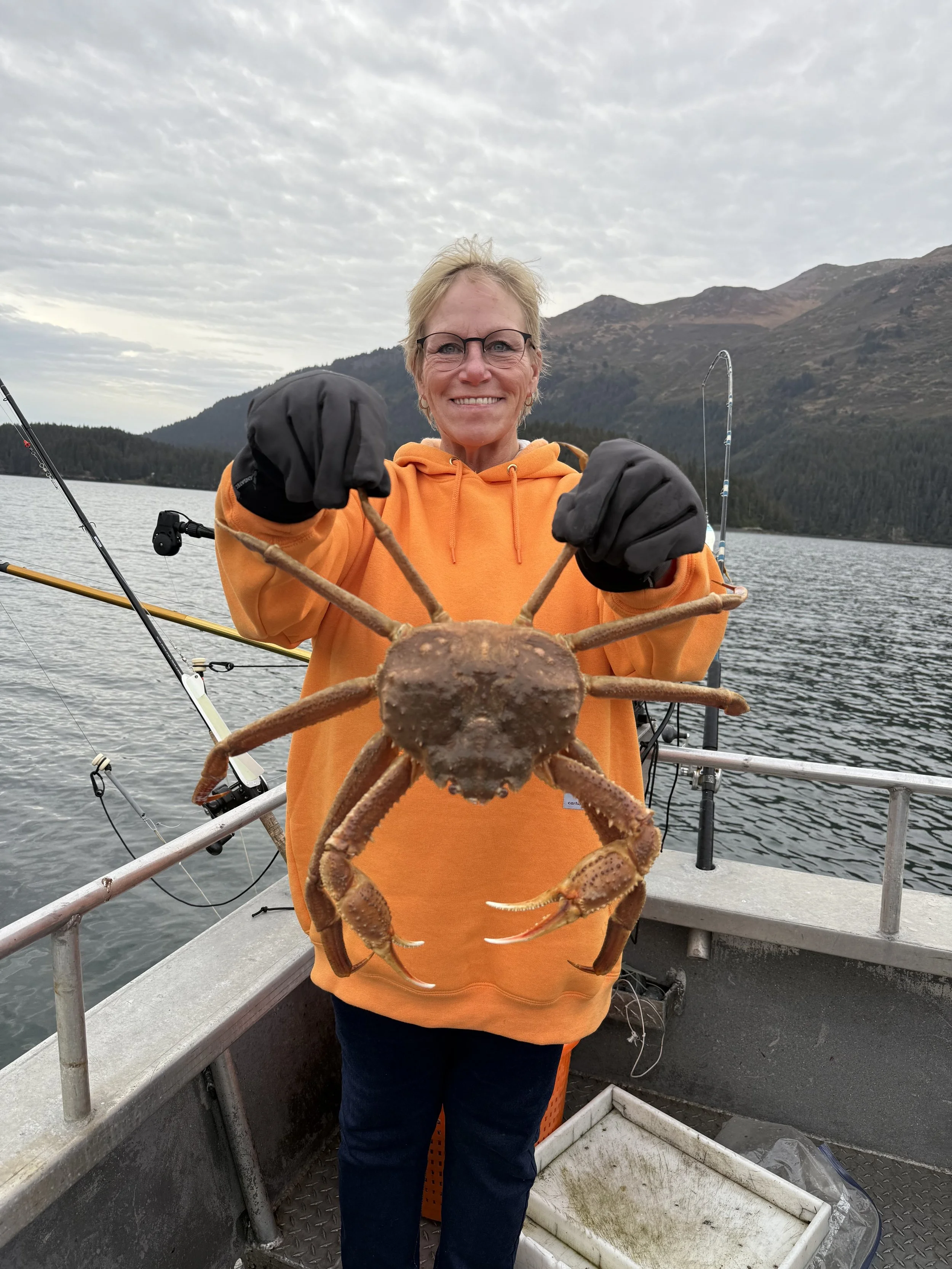 A woman in an orange hoodie holding a large crab on a boat, with mountains and water in the background.