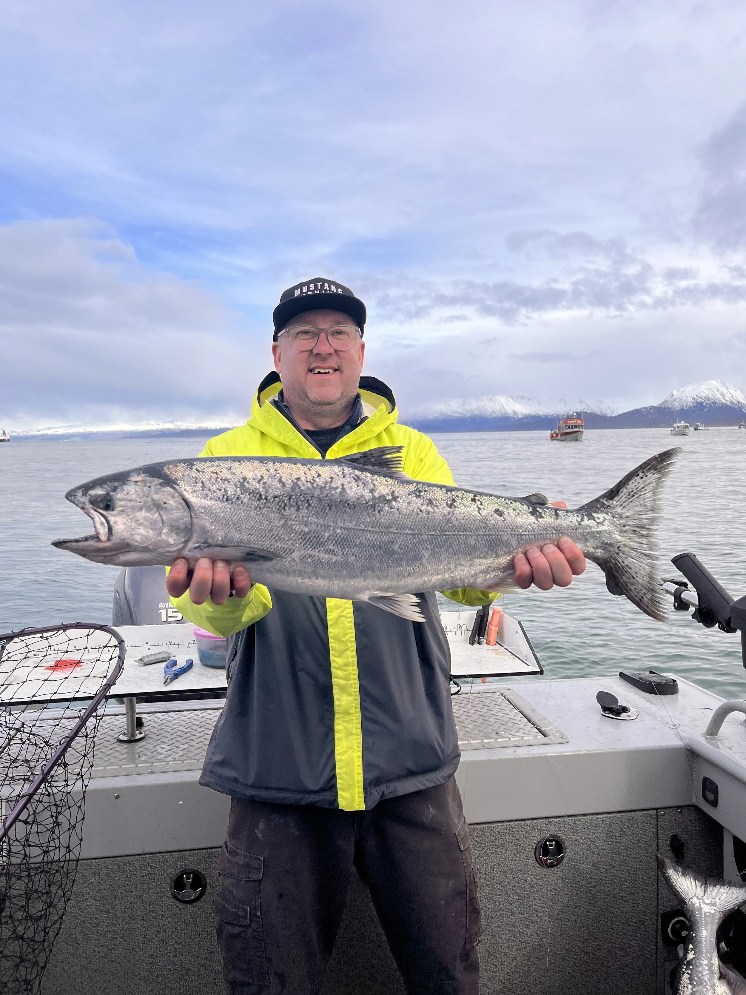 Man in yellow jacket holding large fish on boat with water and snow-capped mountains in background.