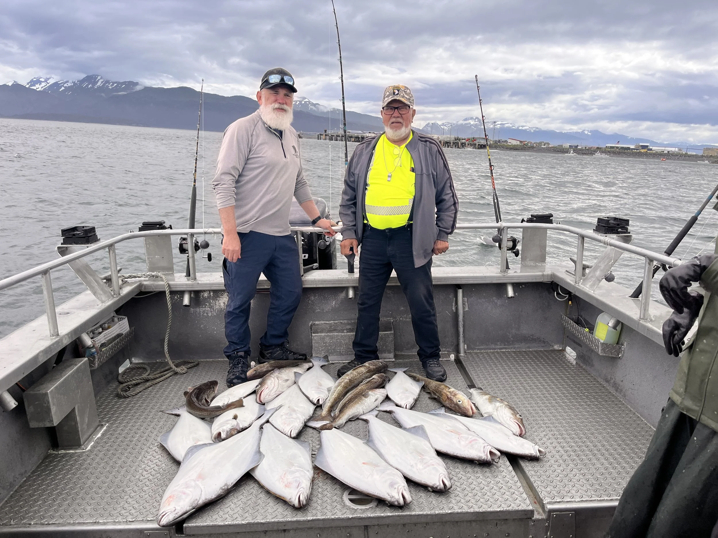 Two men standing on a boat with a catch of fish, including cod and halibut, in a harbor with mountains in the background.